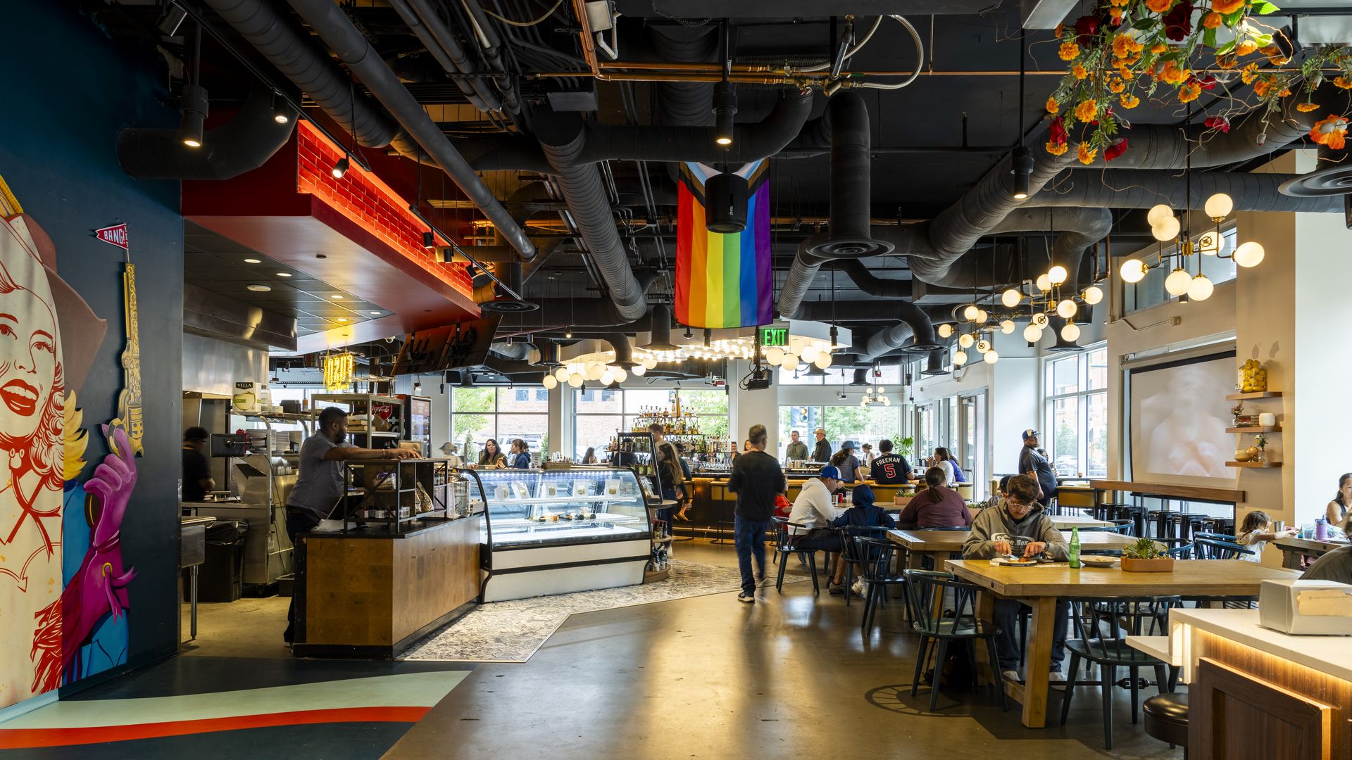 A large food hall with several table, and people sitting and walking. A rainbow flag hangs from a ceiling with exposed tubing.