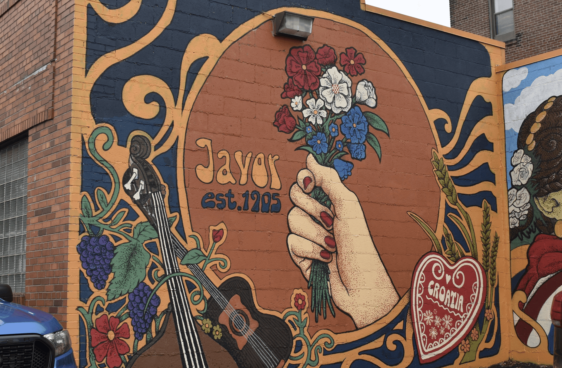 Colorful mural on a brick wall: a hand holding a bouquet of red, white, and blue flowers, a guitar nearby, swirling blue and orange vines, and the text "Taylor est. 1705".