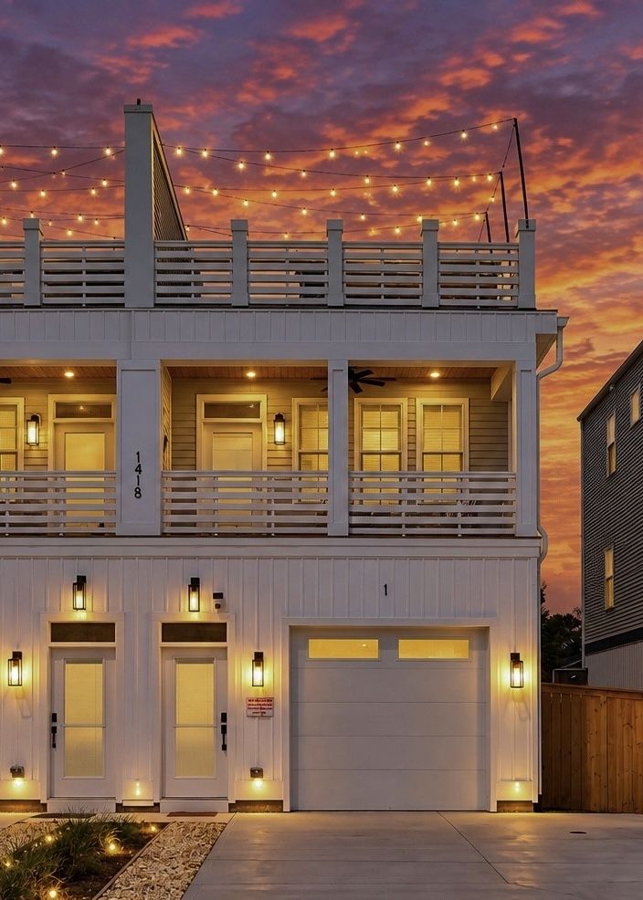 Two-story white home with a garage and upper balcony, string lights across the deck, warm exterior lights at doors and windows, as an orange sunset colors the sky.