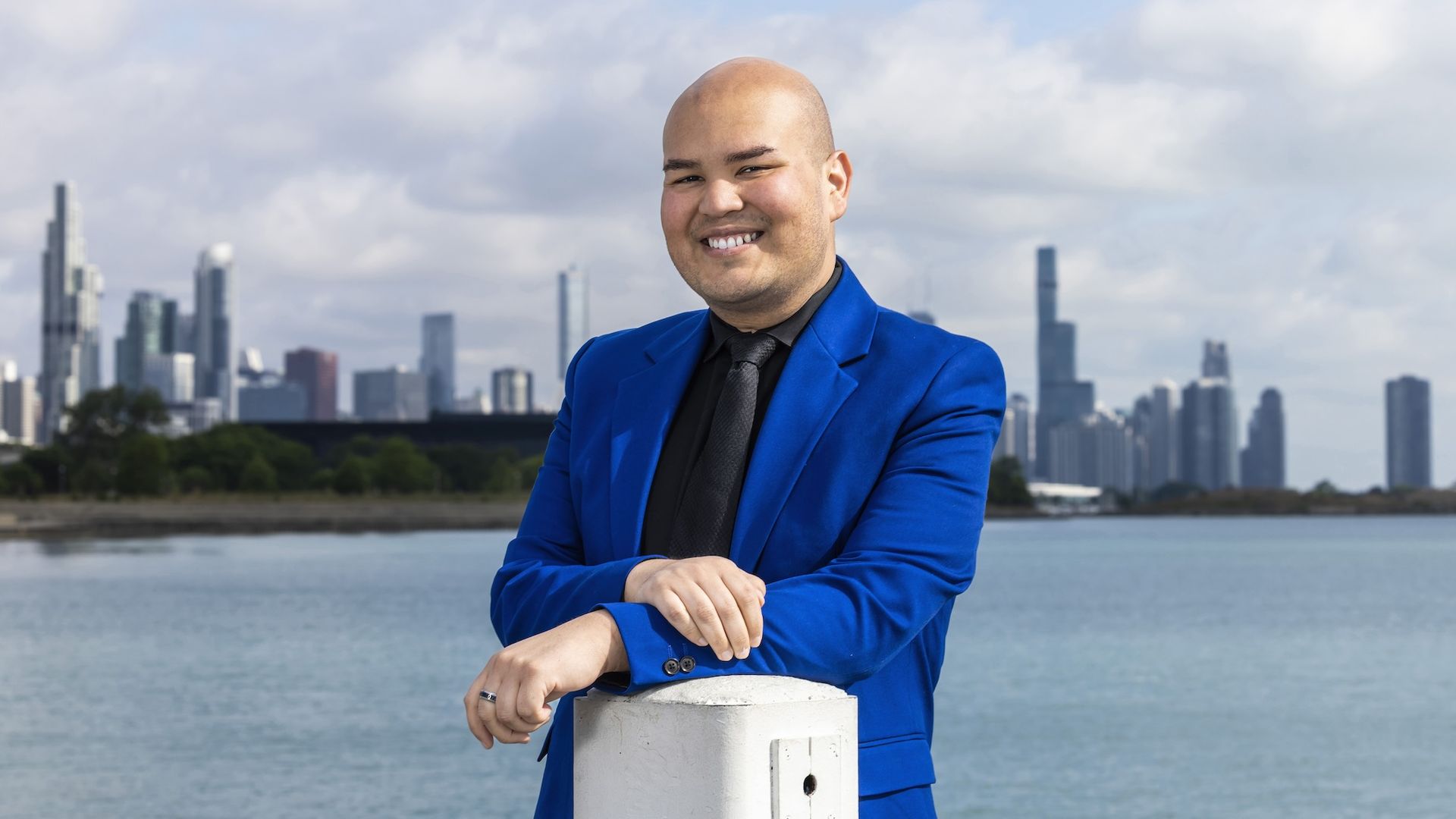 Man in blue suit with black shirt standing in front of lake with skyline in the background.