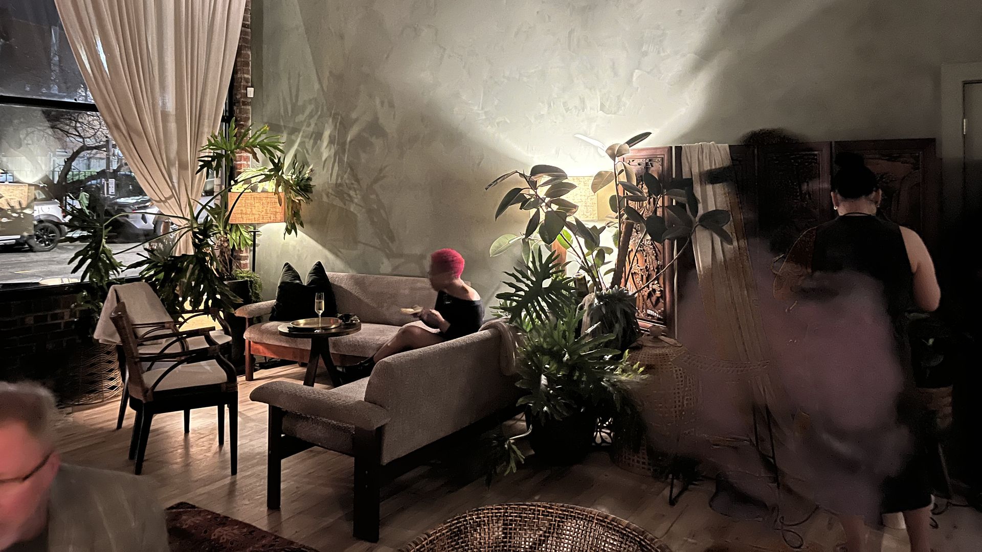 Dimly lit cozy indoor space with plants, beige sofas, wooden chairs, a small table with drinks, and people. Large window with cream curtains shows nighttime outside.