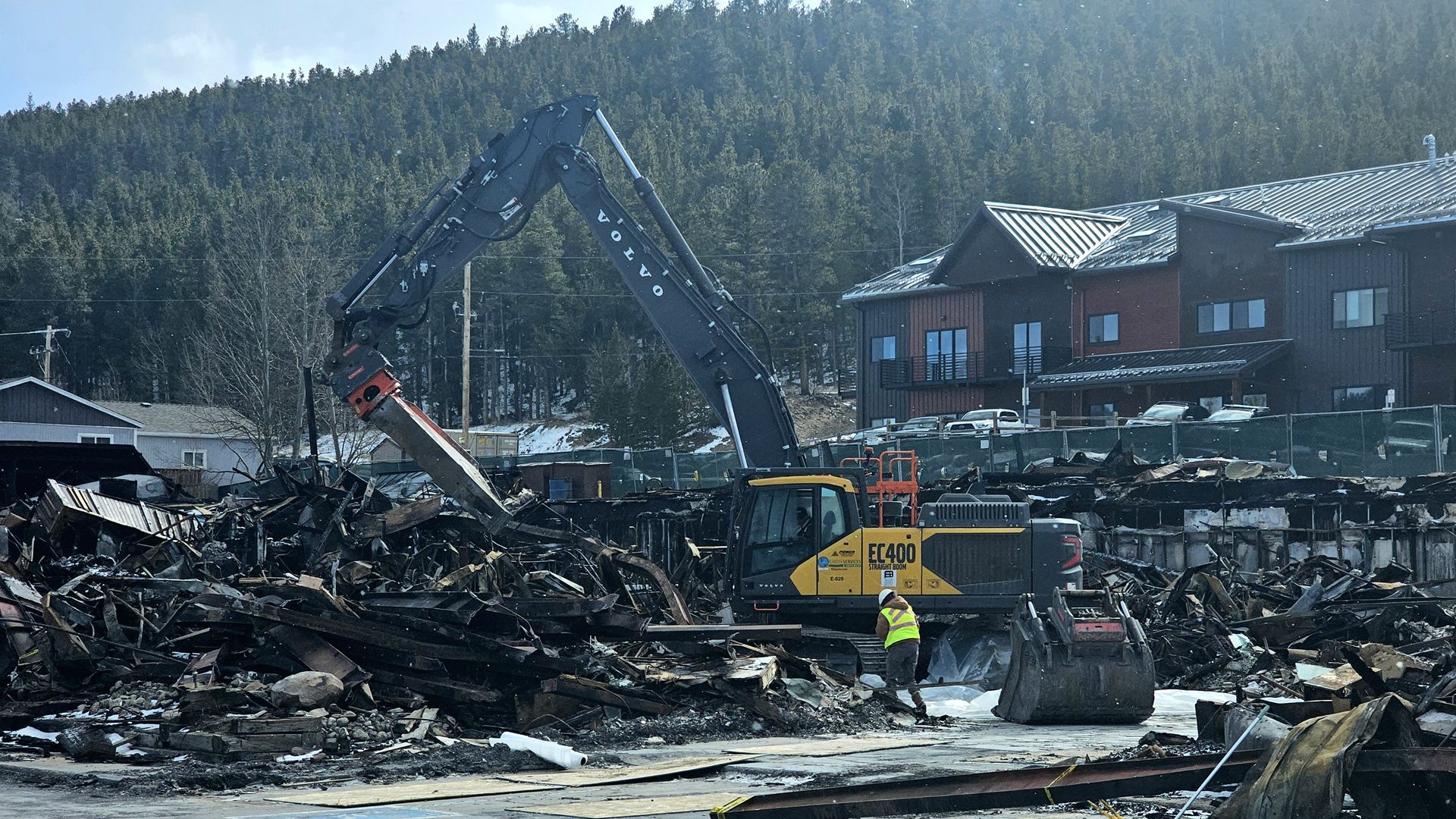 Yellow and black Volvo EC400 excavator clearing charred debris from a fire-damaged site with a worker in a neon vest and helmet nearby, surrounded by forest and modern buildings.