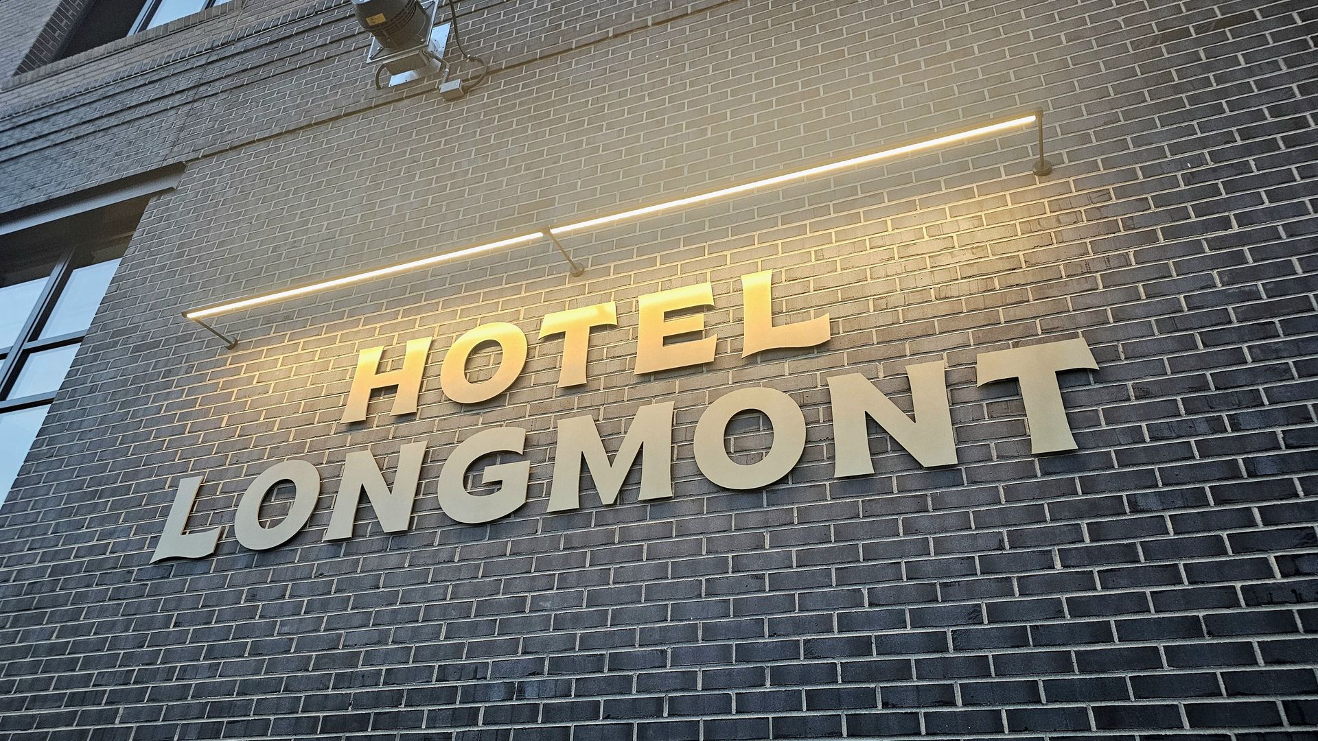 Diagonal view of a gray brick hotel wall with a warm yellow neon strip above large white letters reading 'LONGMONT' and the partial 'HOTEL' text below.