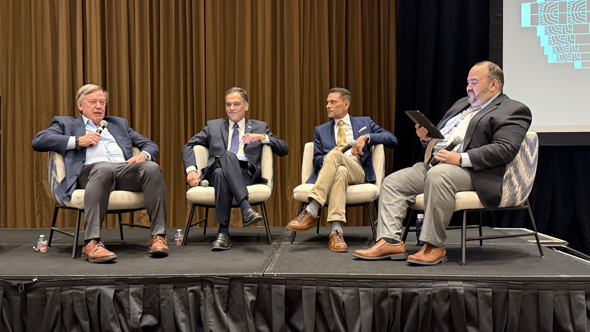 Four men in suits sit on a panel on a stage with brown curtains; microphones in hand, the far-left man speaks into a microphone and the far-right man holds a tablet. A blue geometric projection appears on a screen to the right.