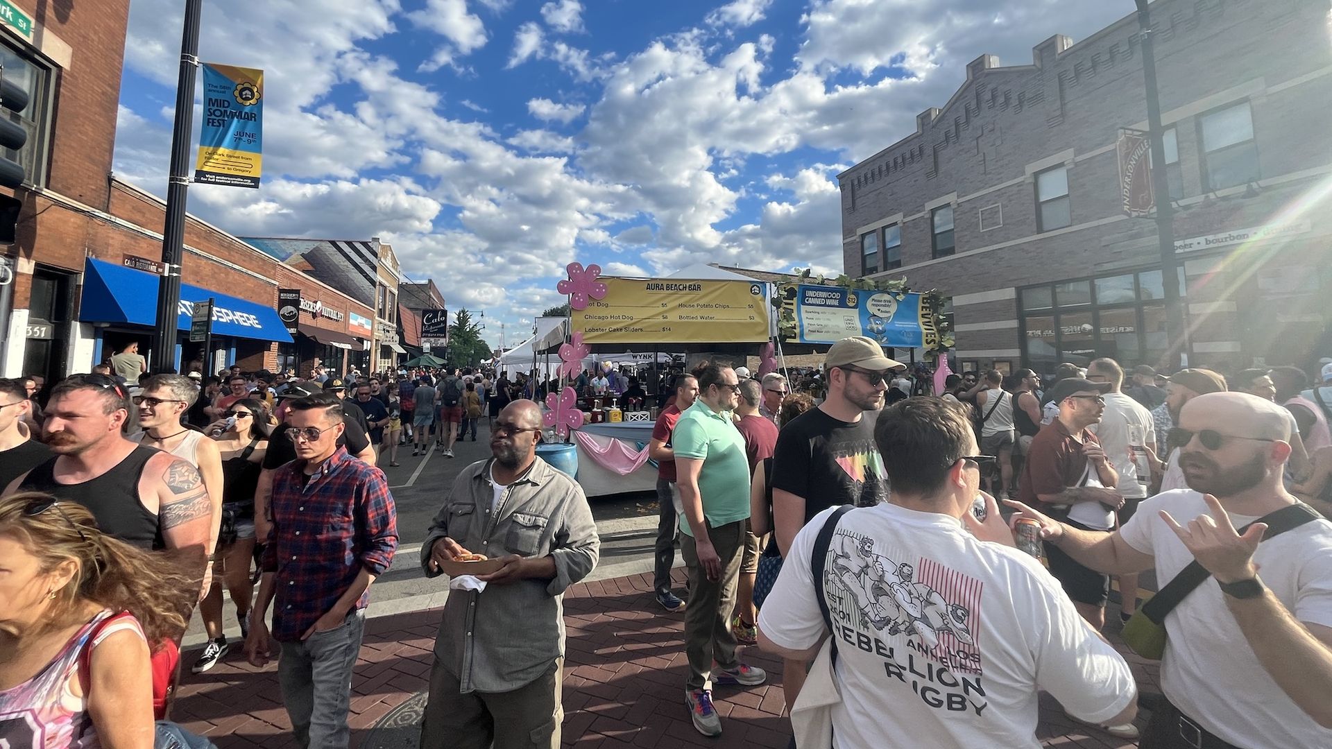 Busy street festival with a large crowd, brick buildings, vendor tents, and colorful banners under a bright blue sky with fluffy clouds.