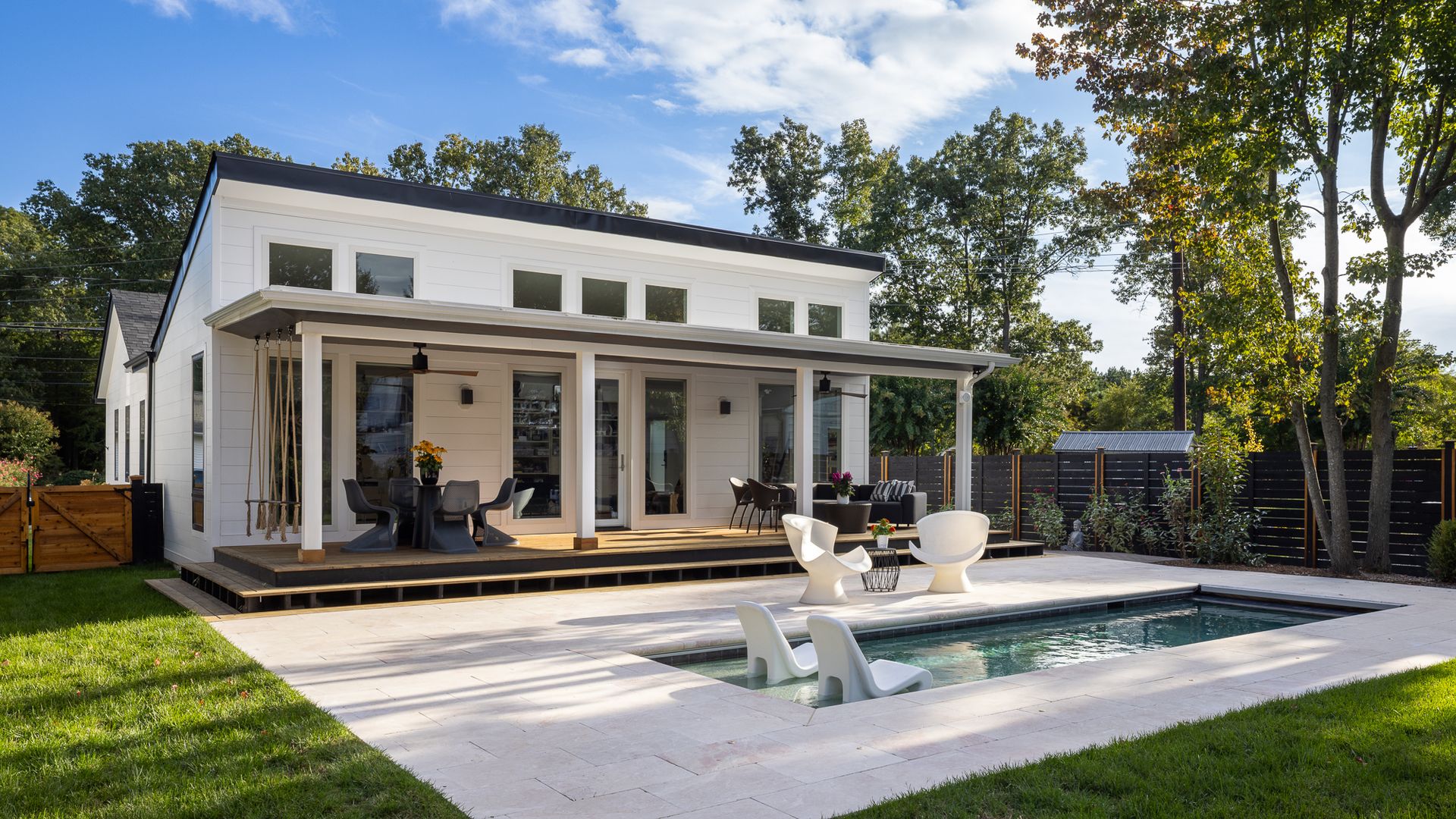 Modern white house with a flat roof and large glass doors opens to a wooden deck beside a rectangular pool. White lounge chairs and circular seating sit on a light tile patio amid lawn and trees.