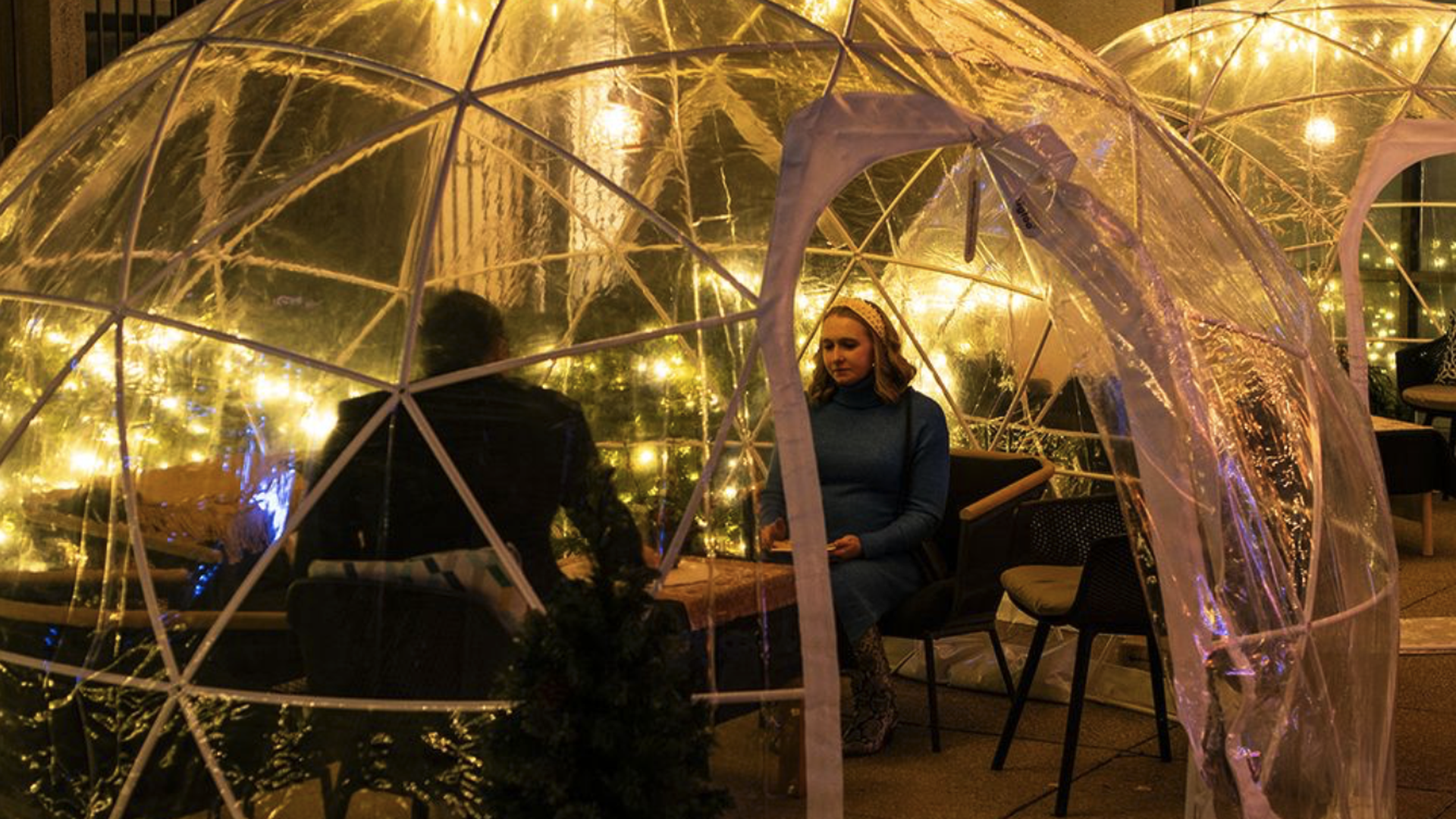 Two people seated inside a transparent, dome-shaped outdoor enclosure decorated with warm yellow string lights at night.