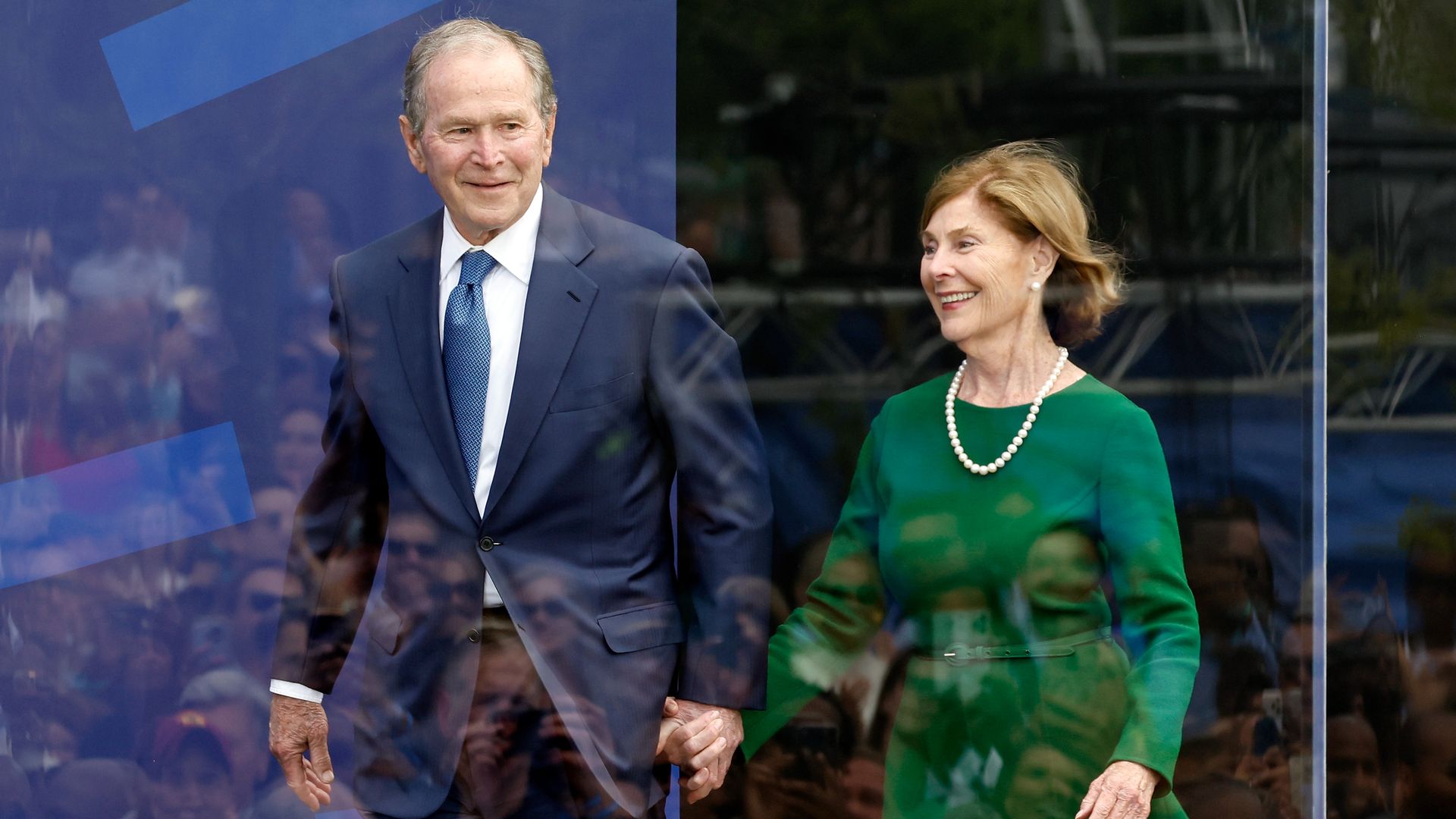 Two elderly people, a man in a navy suit and blue tie and a woman in a green dress with a pearl necklace, smile and stand hand-in-hand behind glass at a formal event.