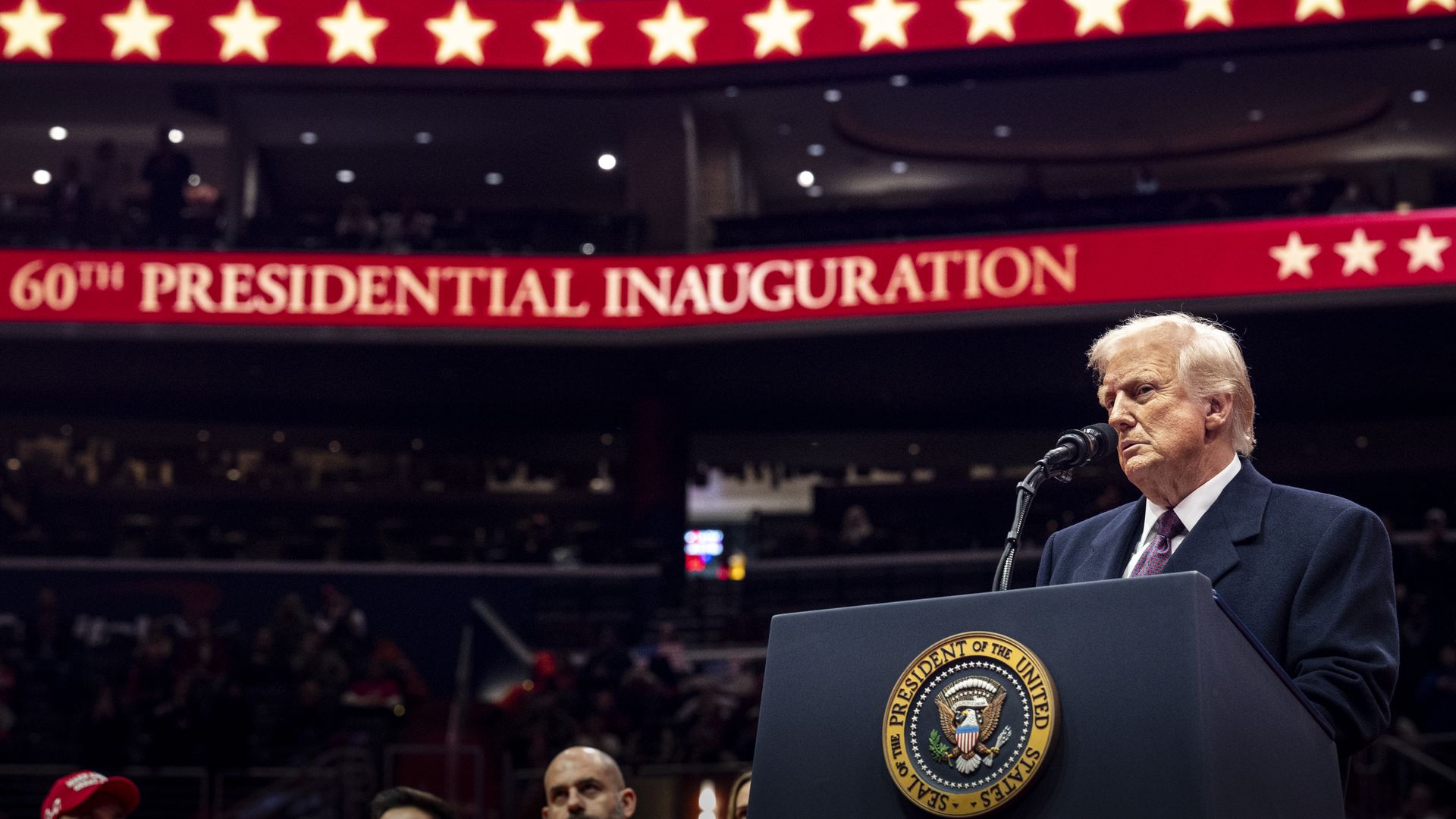 President Trump stands in an arena, behind a podium. Red electronic banners are seen behind him.
