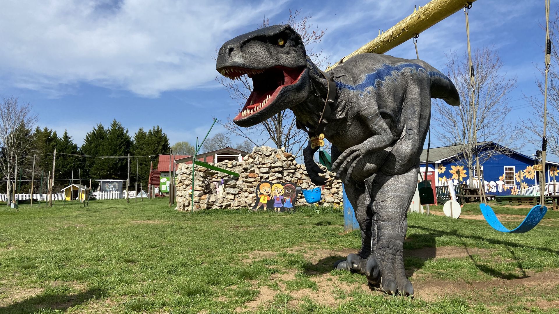 A person wearing a very realistic T. Rex costume walks underneath a swing set on a partly cloudy but pretty day.