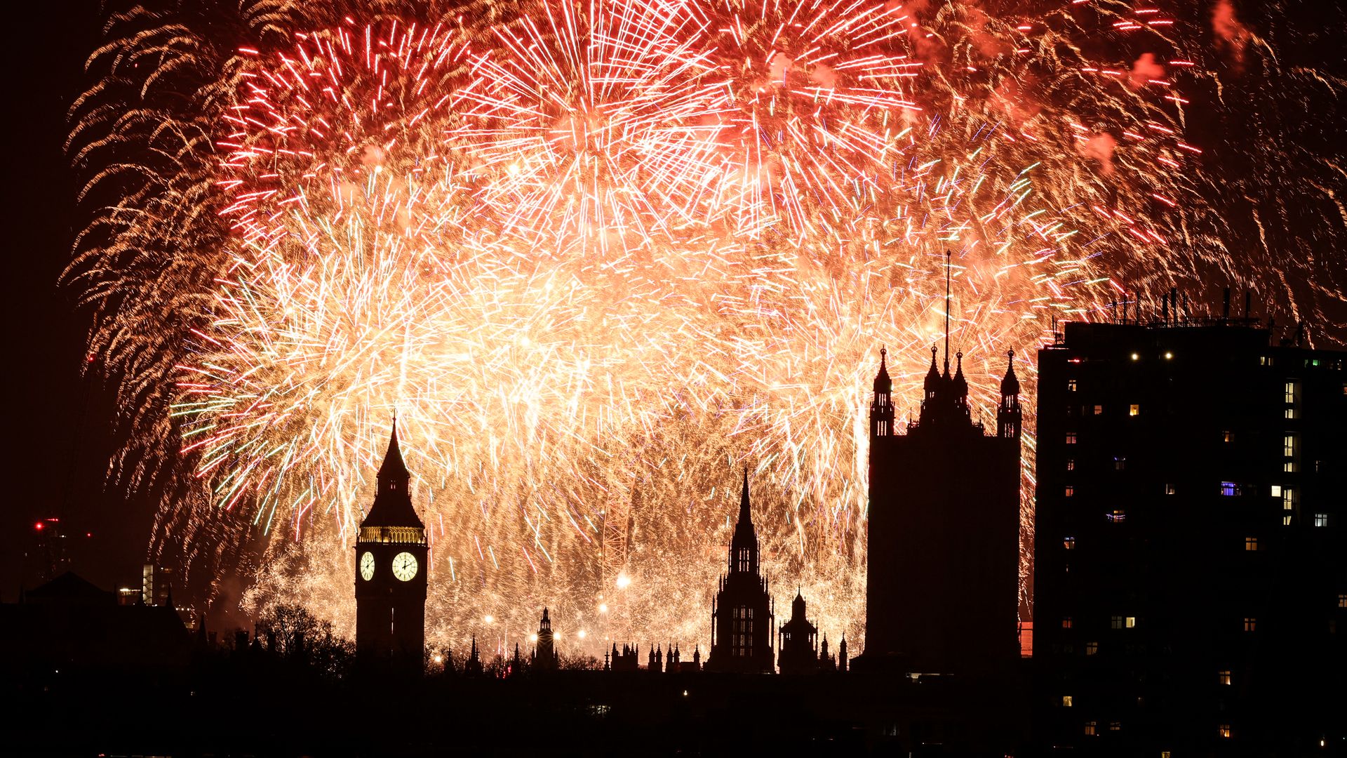 Bright red and gold fireworks exploding in the night sky above the silhouette of London landmarks, including Big Ben, with a dark building on the right side lit by small windows.