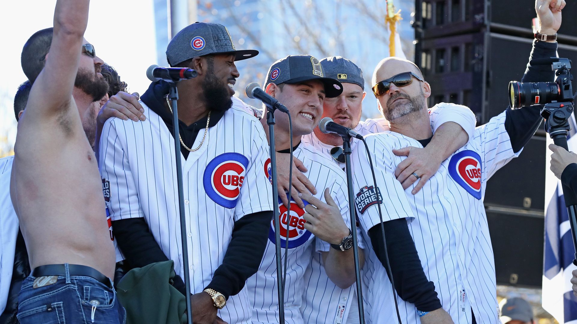 Group of Chicago Cubs players in white pinstripe jerseys and hats celebrating on stage with microphones, one shirtless man raising his arm, and a cameraman filming the scene outdoors.
