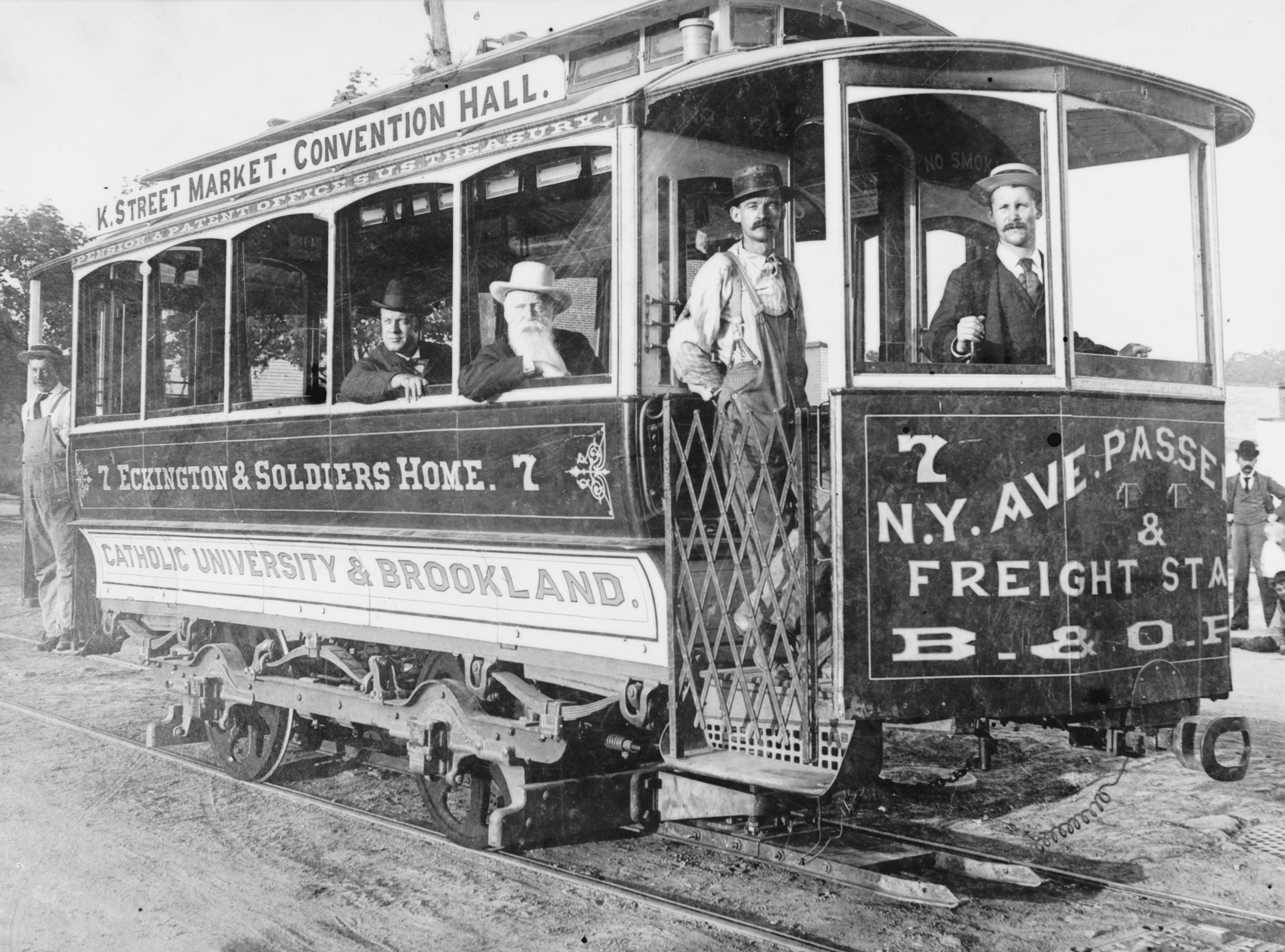 Black-and-white photo of a vintage streetcar with open doors and several men inside and on the steps. Signs read K Street Market, Convention Hall, Catholic University & Brookland, and N.Y. Ave & Freight Sta.