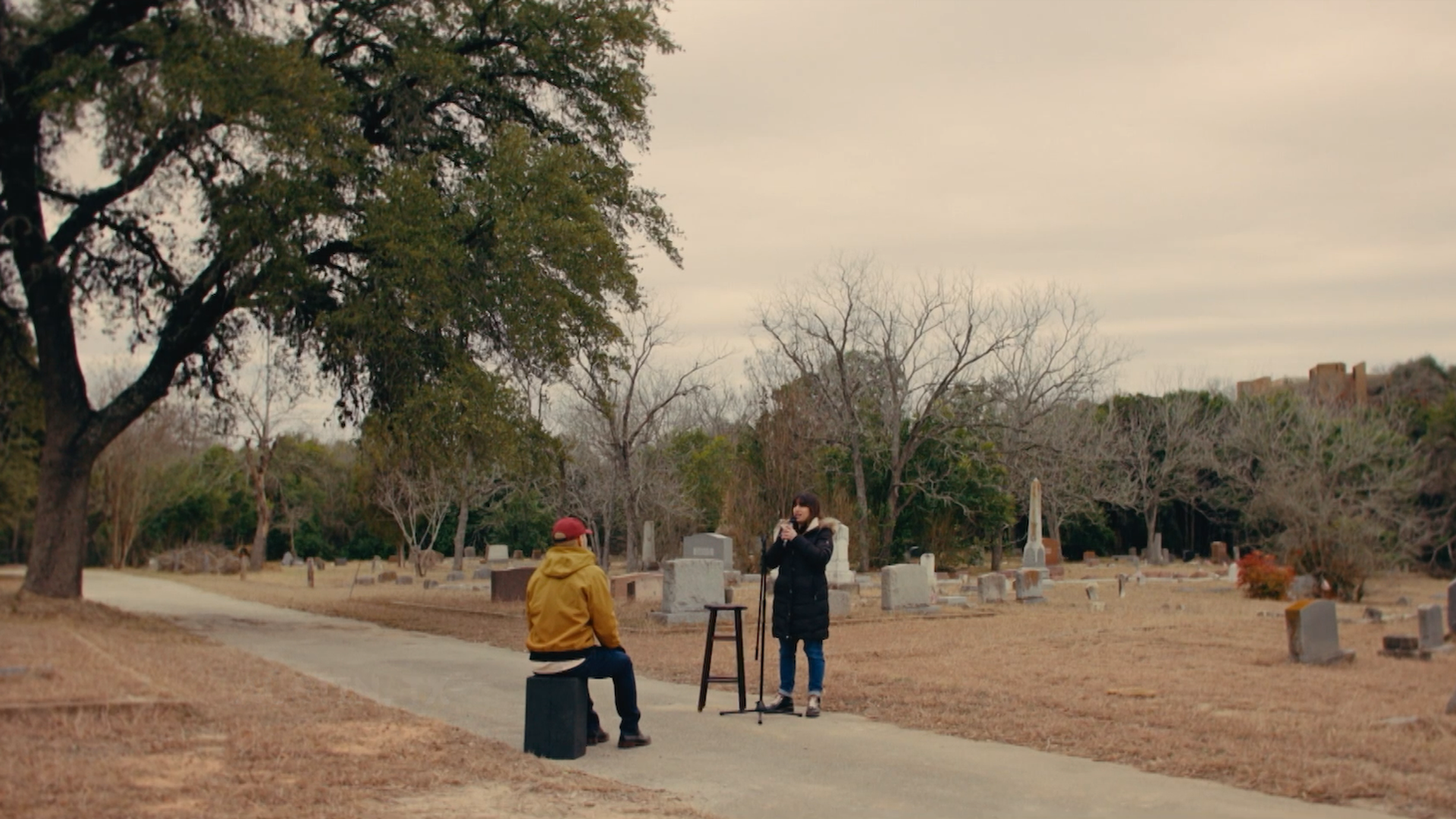 Two people in a cemetery: one in a yellow jacket and red cap sits on block by path, while another in a dark coat speaks into a microphone beside a tripod; leafless trees and tombstones under pale sky.