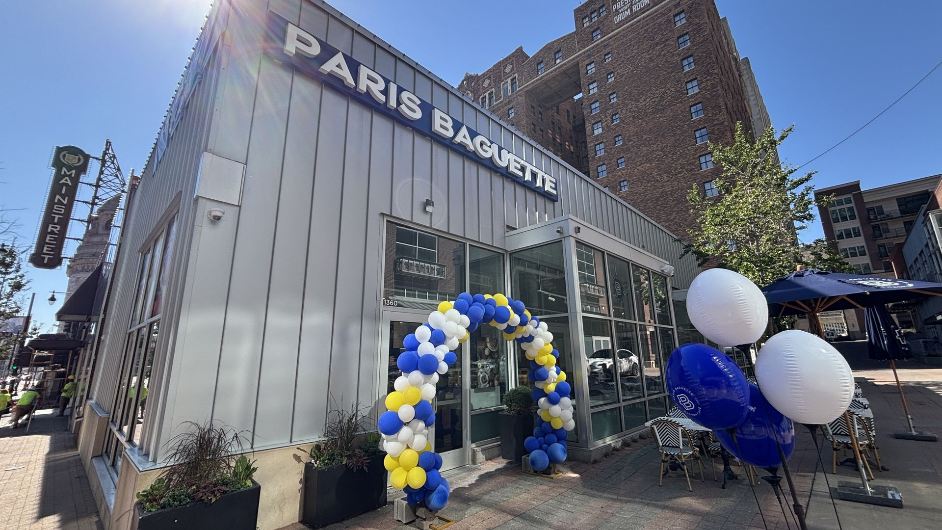 Paris Baguette bakery on Main Street with a glass storefront, a blue, white and yellow balloon arch, and large white/blue balloons outside; tall brick building in the background on a sunny day.