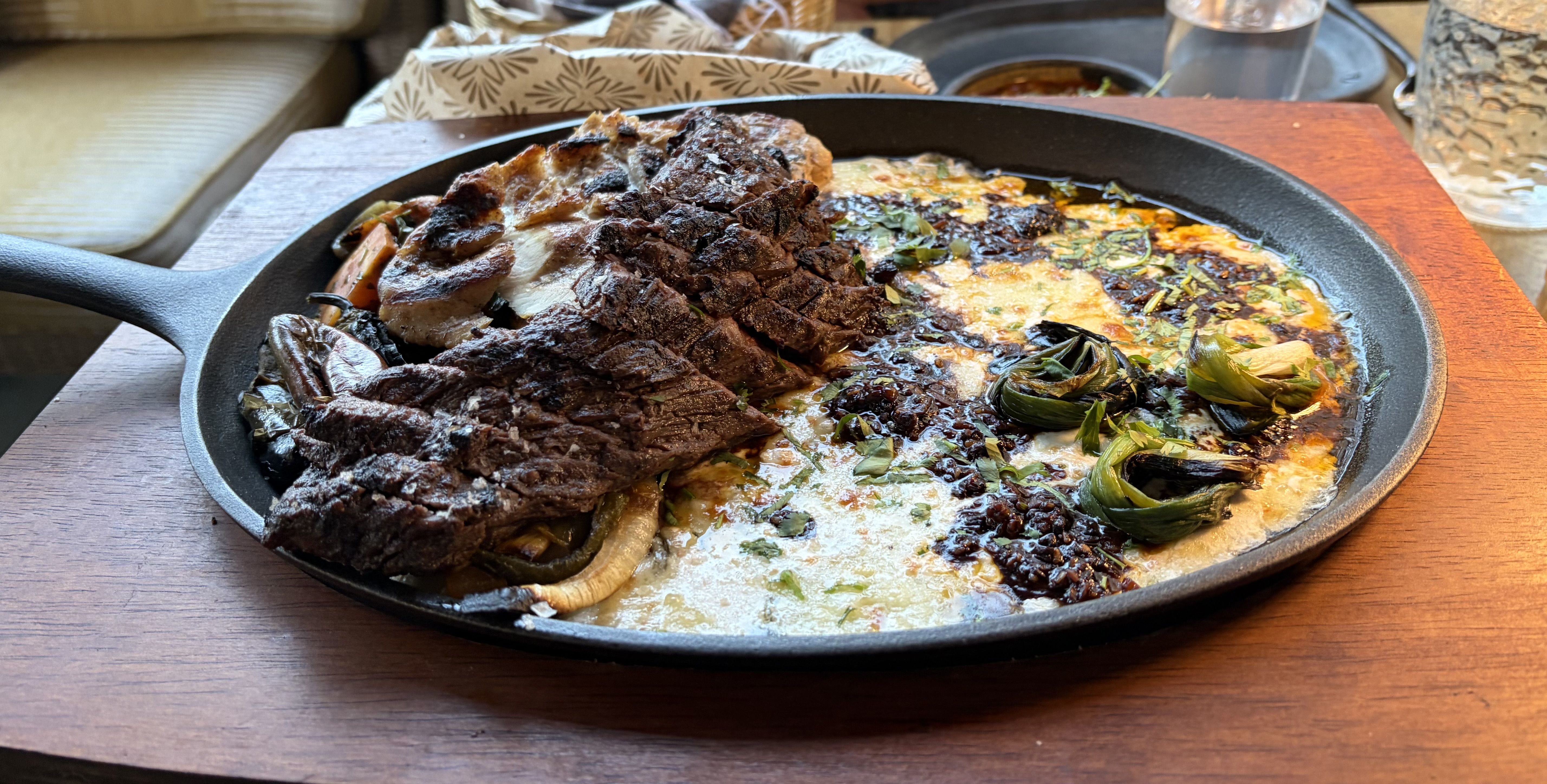 Cast-iron skillet on a wooden table with thick, charred beef slices over sautéed onions and greens, plus mushrooms and melted cheese, garnished with herbs.