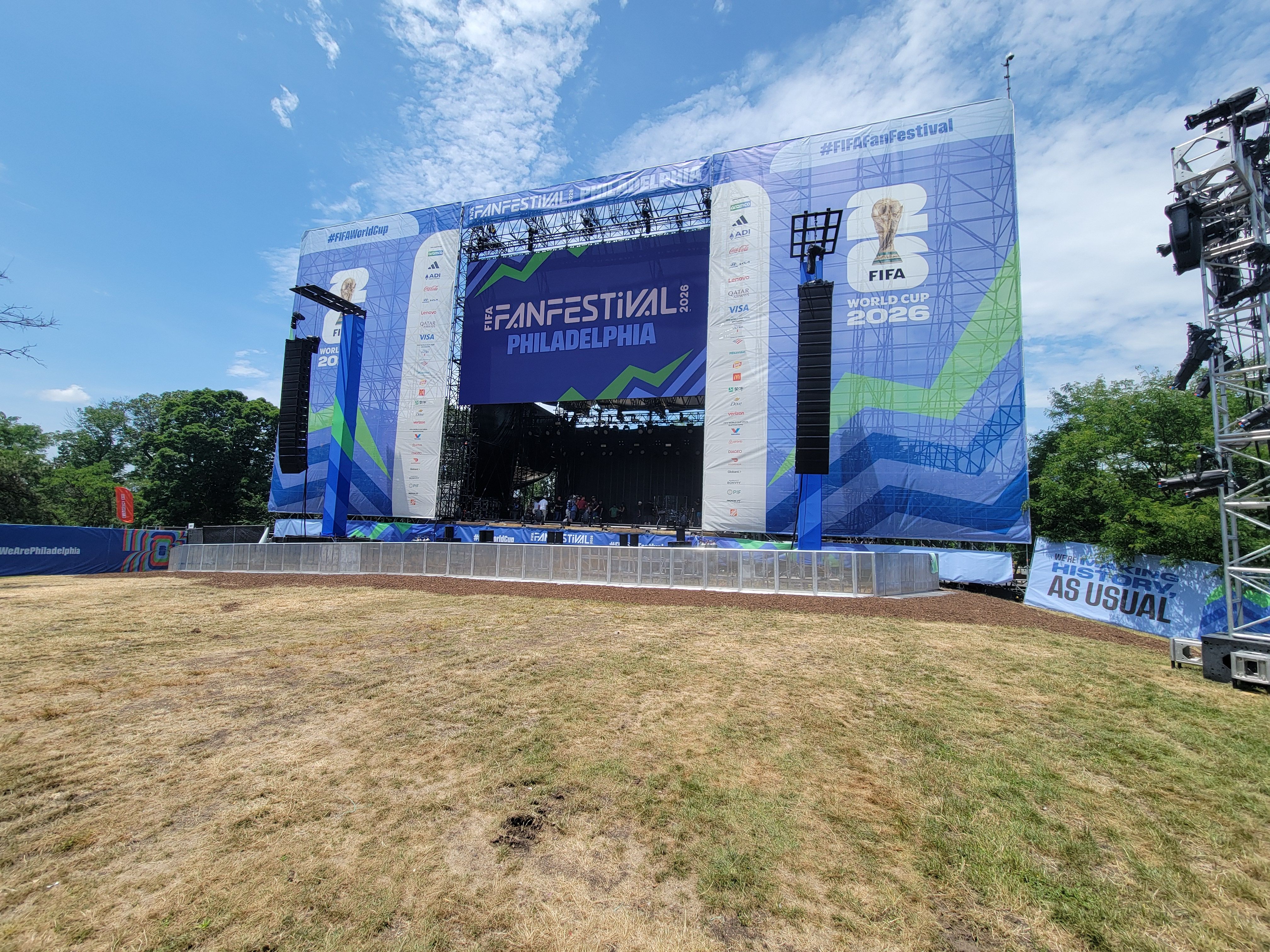 Outdoor festival stage with large blue banners reading FIFA FAN FESTIVAL Philadelphia and World Cup 2026, tall speakers, metal scaffolding, and a bright blue sky with white clouds.