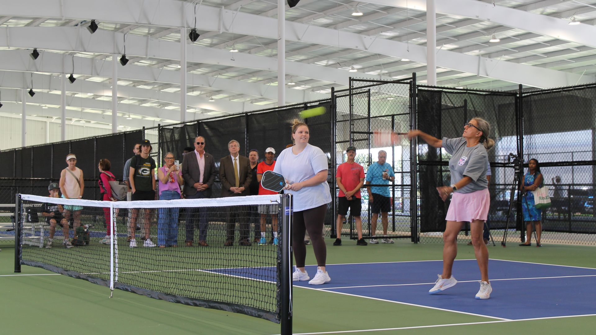 Indoor tennis facility with two female players near the net on a blue and green court, while a crowd watches from behind a chain-link fence under bright white warehouse lights.