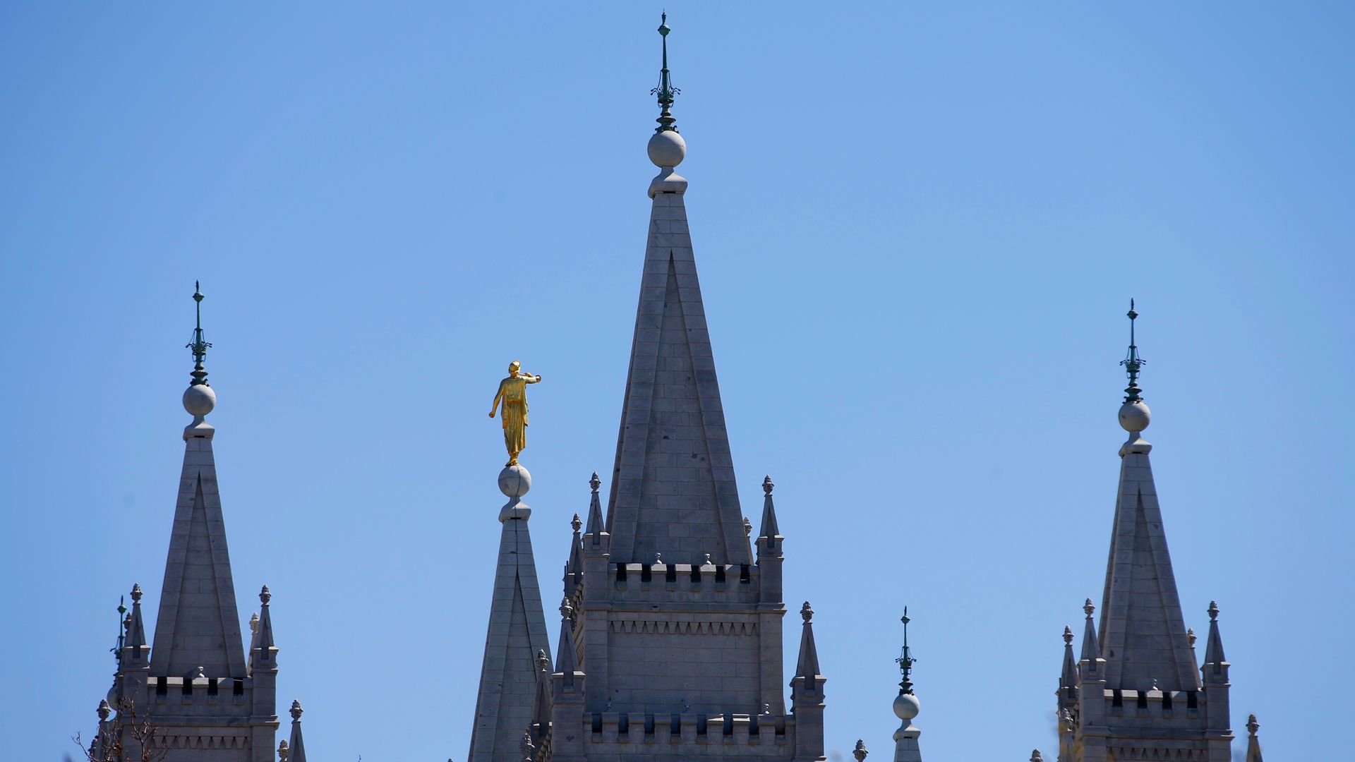 The spires of the Salt Lake City temple of the Church of Jesus Christ of Latter-day Saints appear in front of a blue sky. 