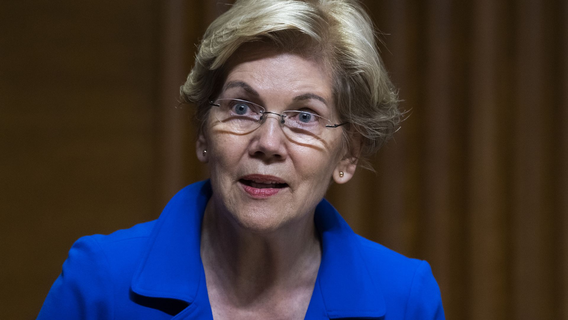A mature woman with short hair, glasses and a blue suit, speaking with a placard in front of her and curtains behind her.