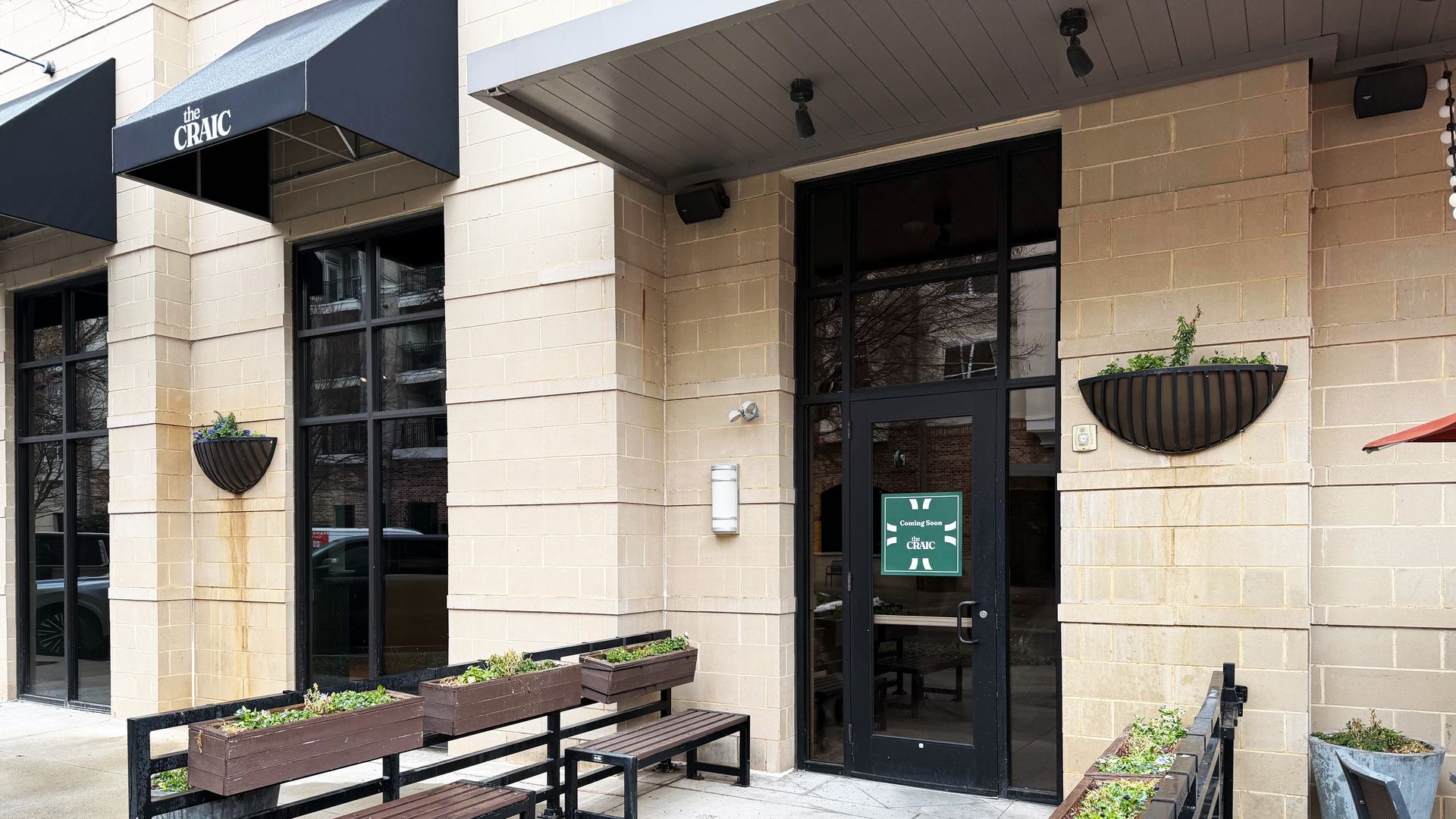 Exterior of a beige brick building with black-framed windows and door featuring a green "Coming Soon the CRAIC" sign, black awnings, wooden benches, and planters with greenery.