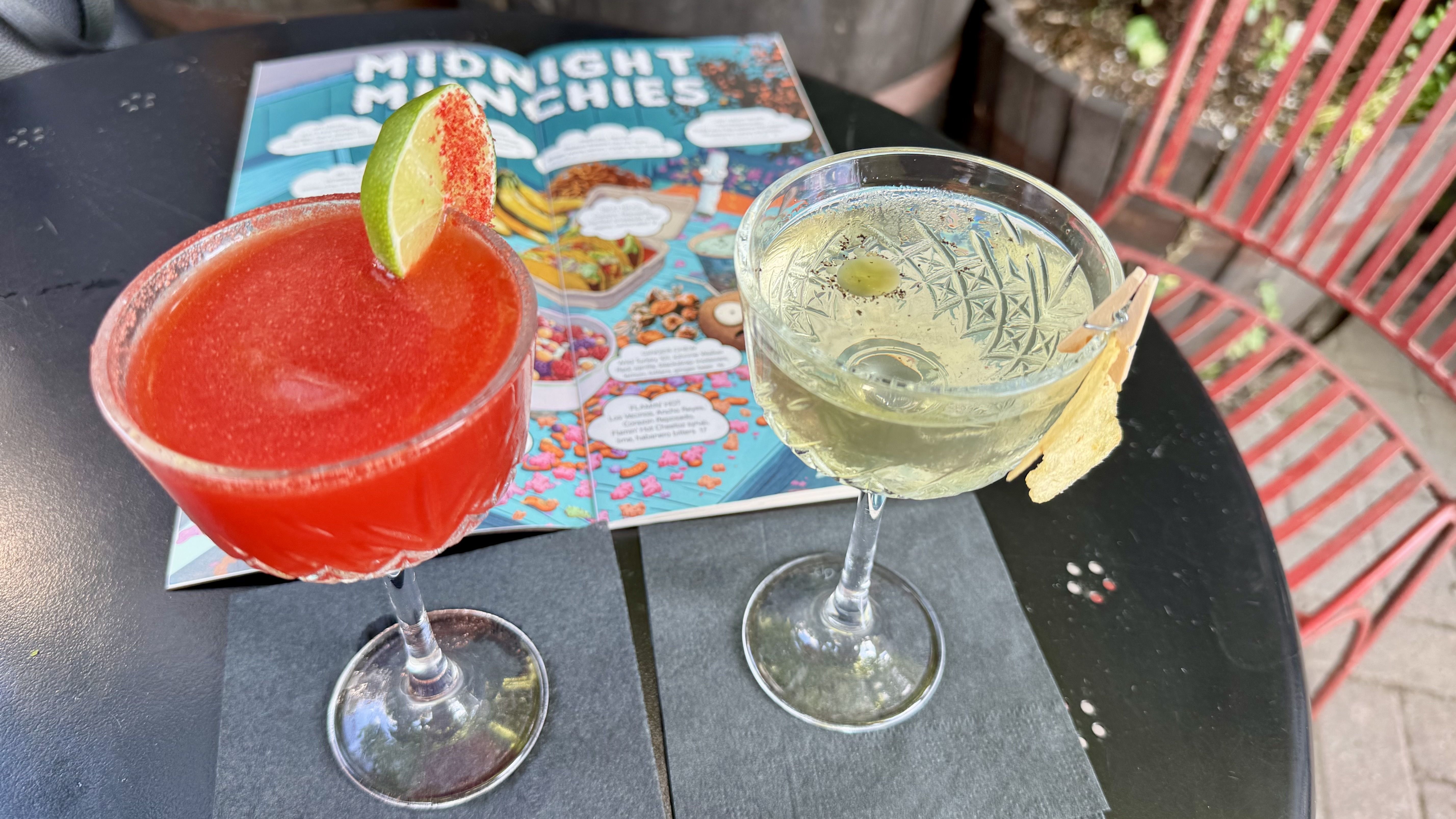 Two cocktails on a black table: a red frozen drink with lime wedge and red rim, and a clear glass with lime and a tortilla-chip garnish; behind is a colorful menu titled "Midnight Munchies".