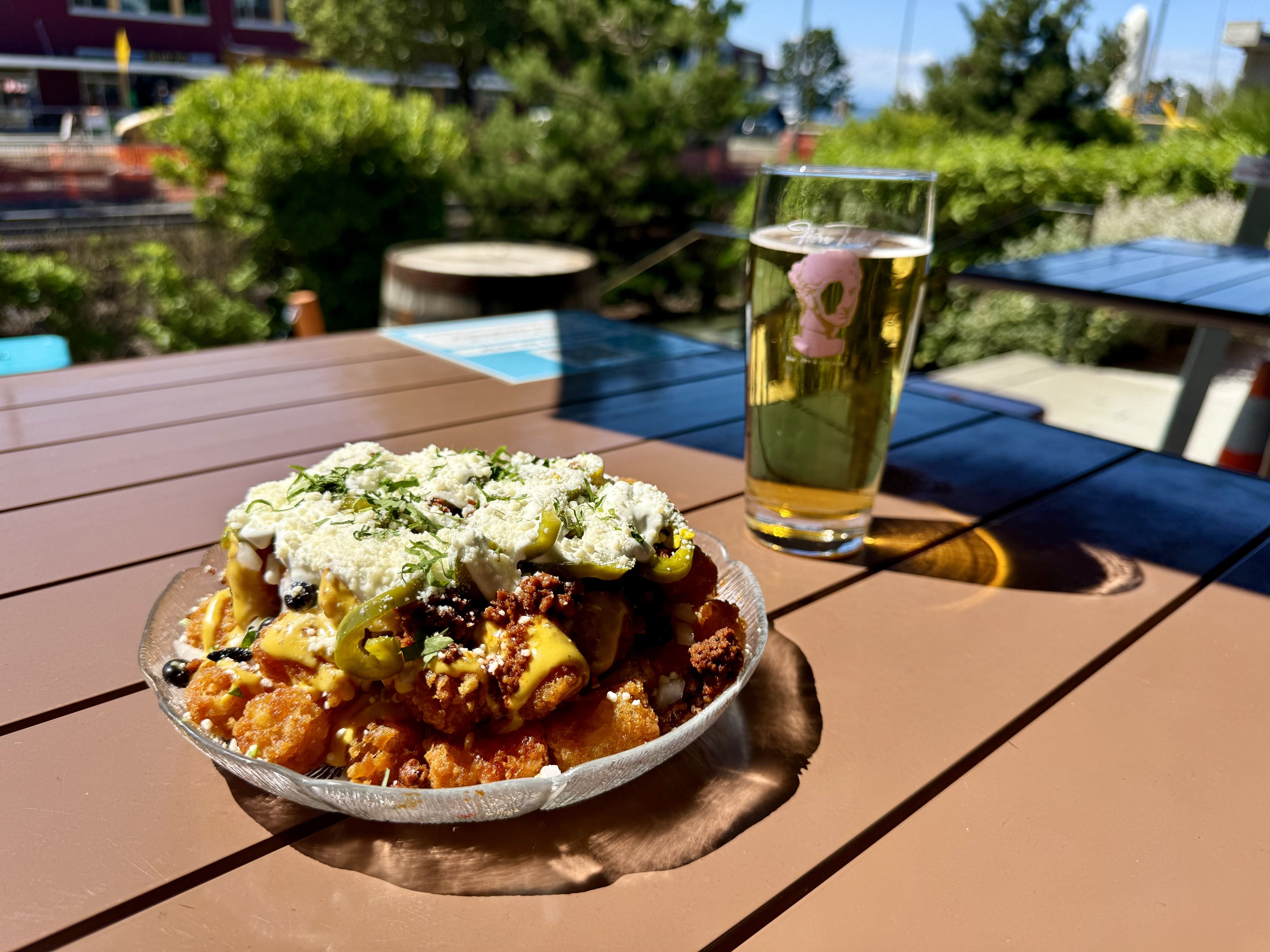 Plate of loaded tater tots topped with cheese, ground meat, jalapeños, sour cream, and herbs on a brown table next to a glass of light beer outdoors on a sunny day.