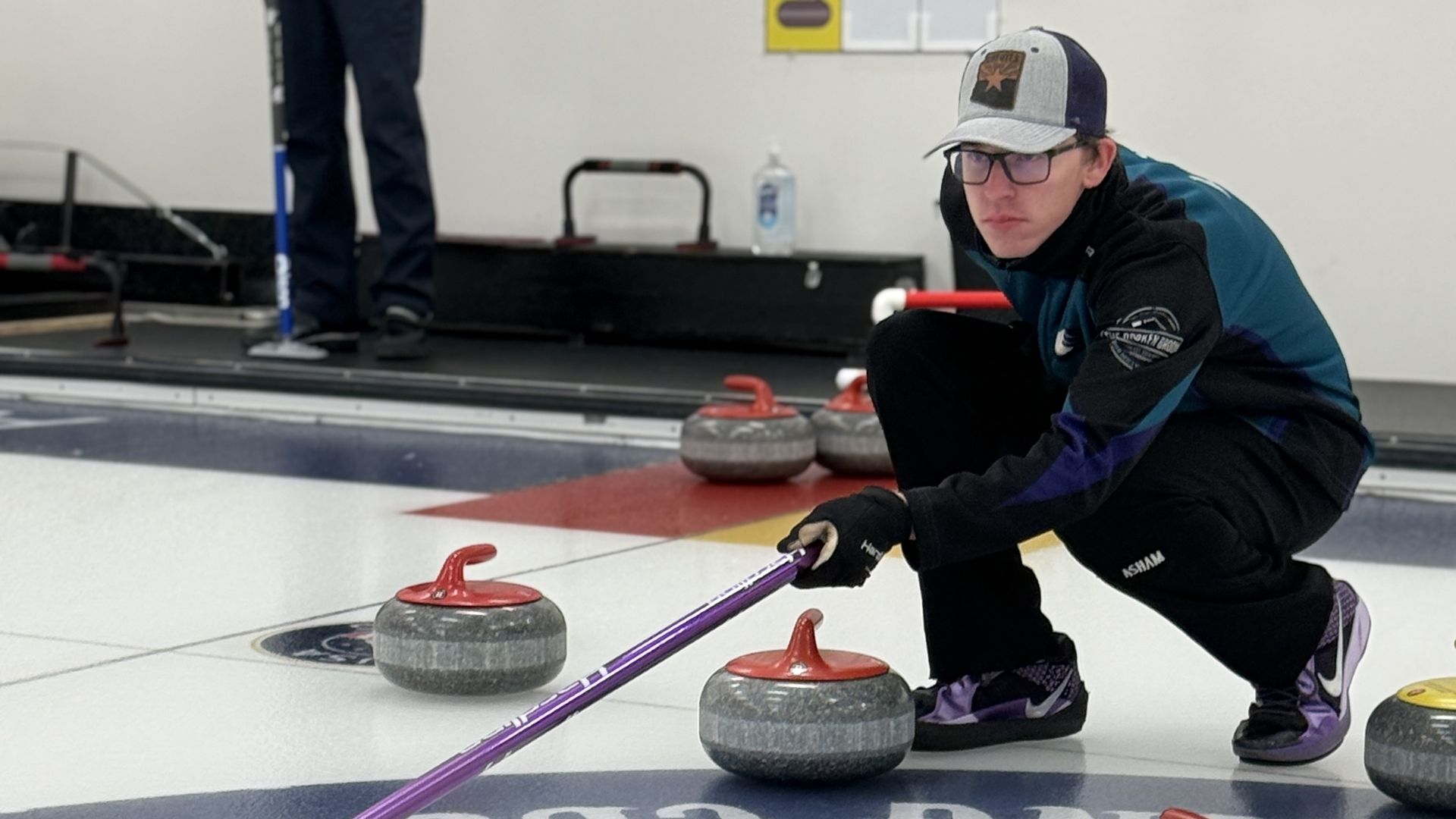 A young man in a gray cap, glasses, and dark sportswear crouches on the curling ice holding a purple broom, surrounded by curling stones with red and yellow handles near the target circle.
