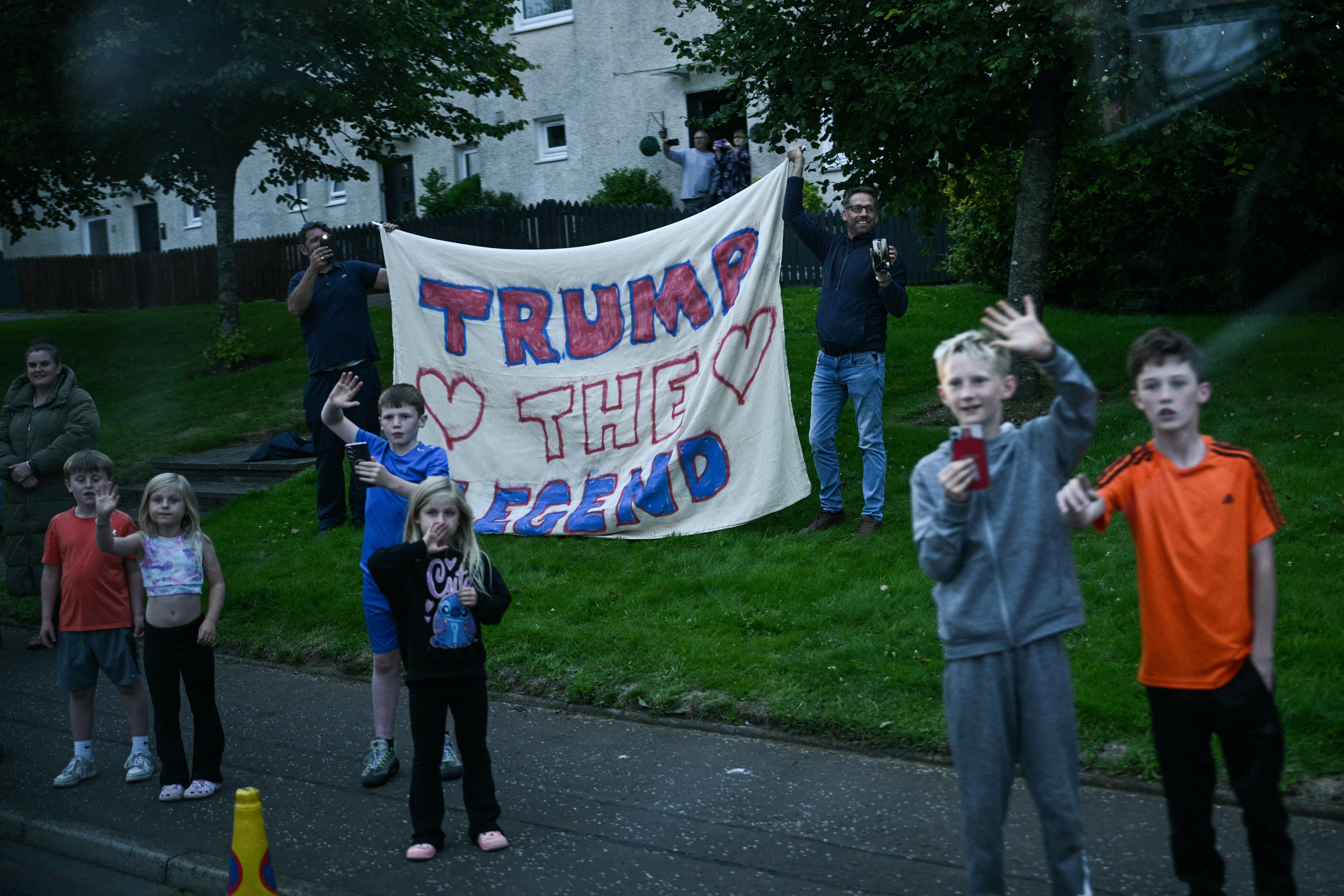 Two men hold a banner reading "Trump The Legend" next to children waving as the US president Donald Trump cars delegation drive past them near Prestwick airport, south of Glasgow on July 25, 2025, on the first day of his visit in the UK.