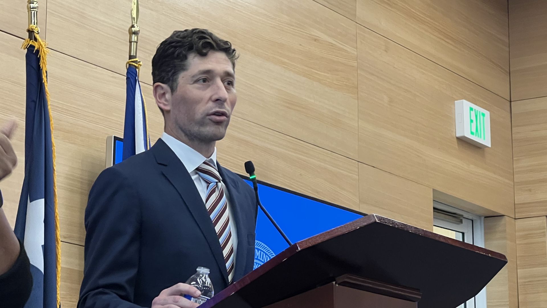 Man in dark suit with striped tie speaking at a podium with the seal of Minneapolis, two flags behind him, a person signing on the left, exit sign on wooden wall