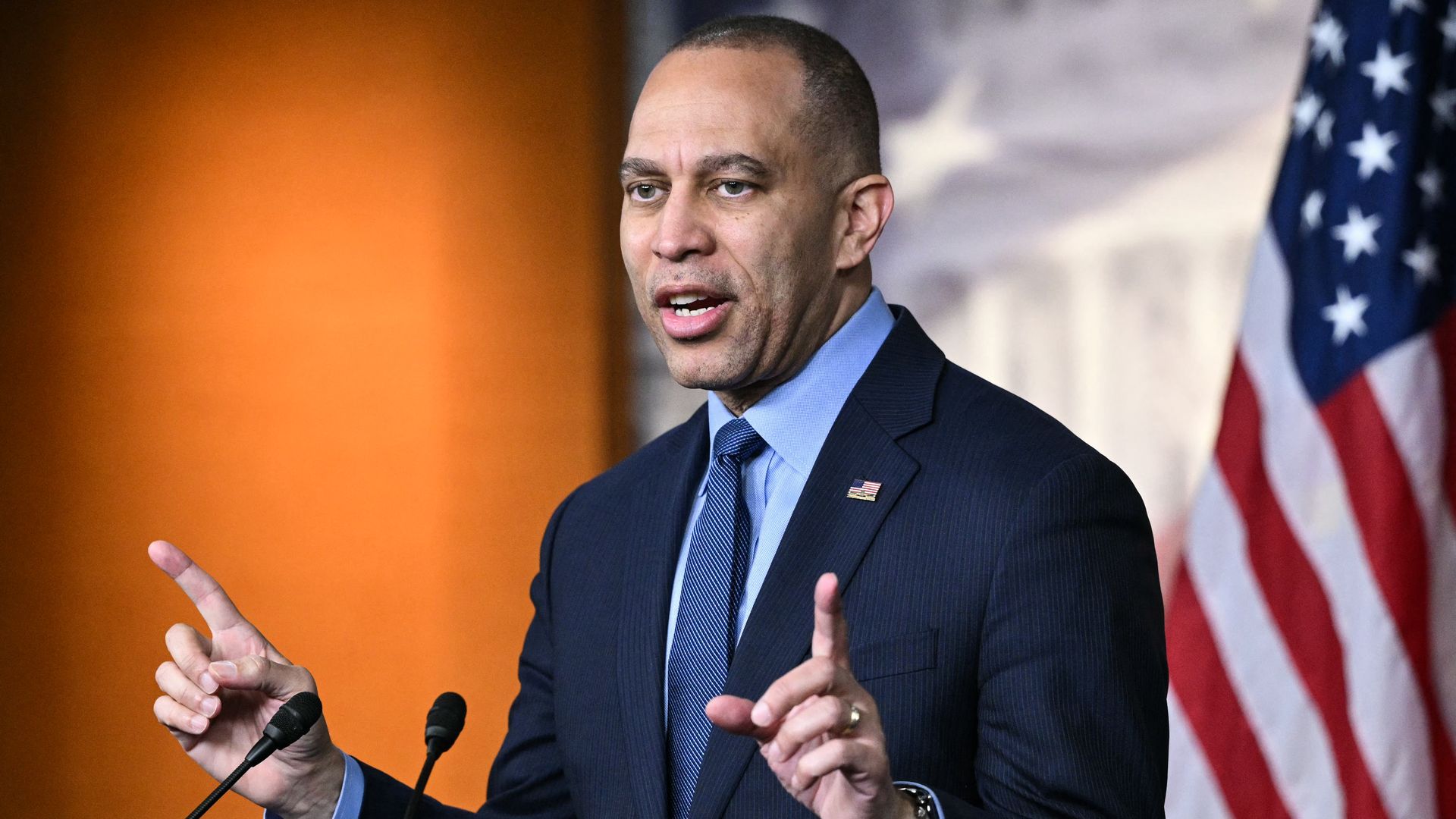 A man in a dark suit and blue tie speaks at a podium with two microphones, gesturing with his hands, with an American flag and an orange backdrop behind him.
