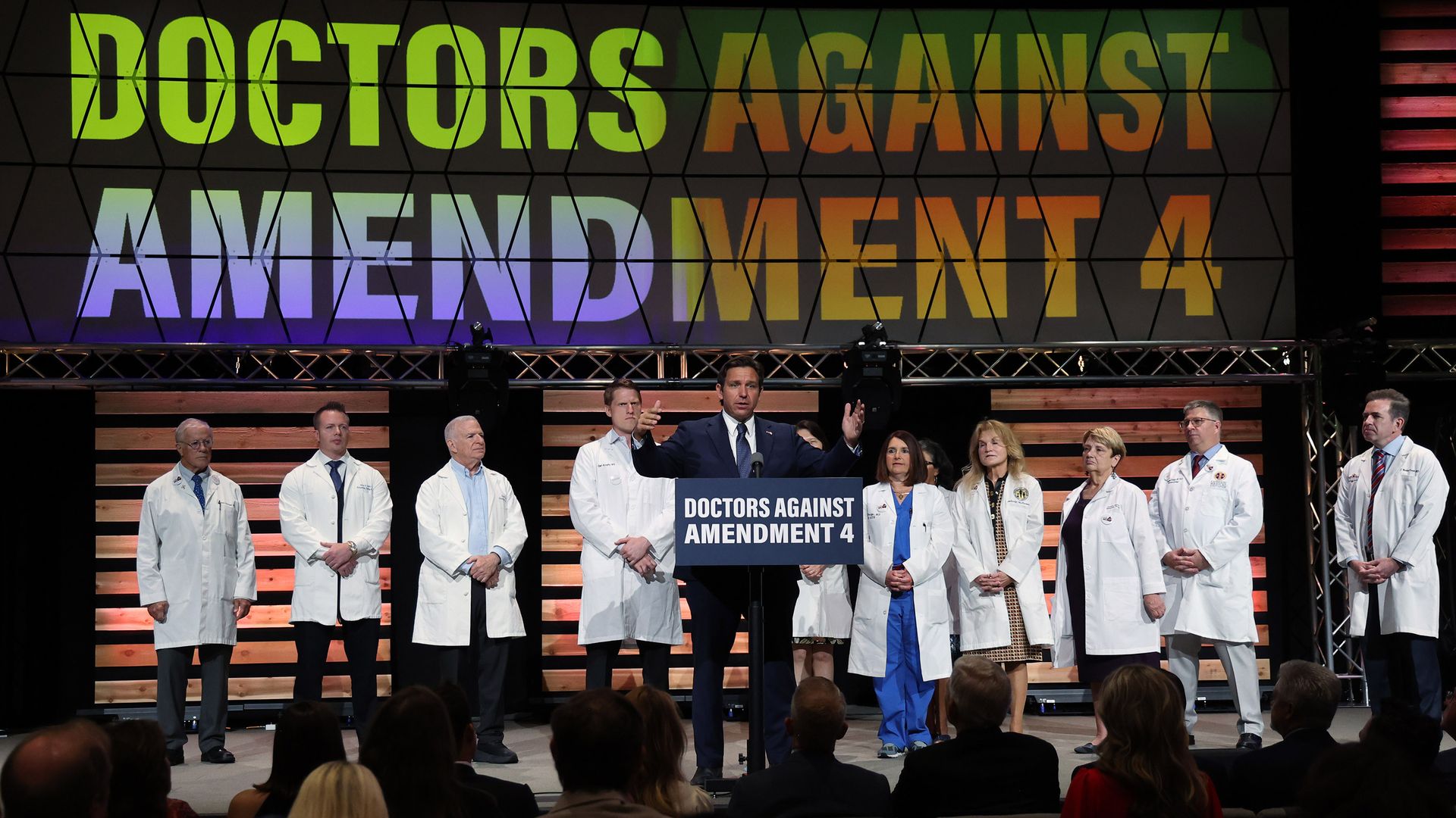 A man in a navy suit stands at a podium that says "DOCTORS AGAINST AMENDMENT 4." Behind him are 10 people in white lab coats. A banner above them reads "DOCTORS AGAINST AMENDMENT 4."