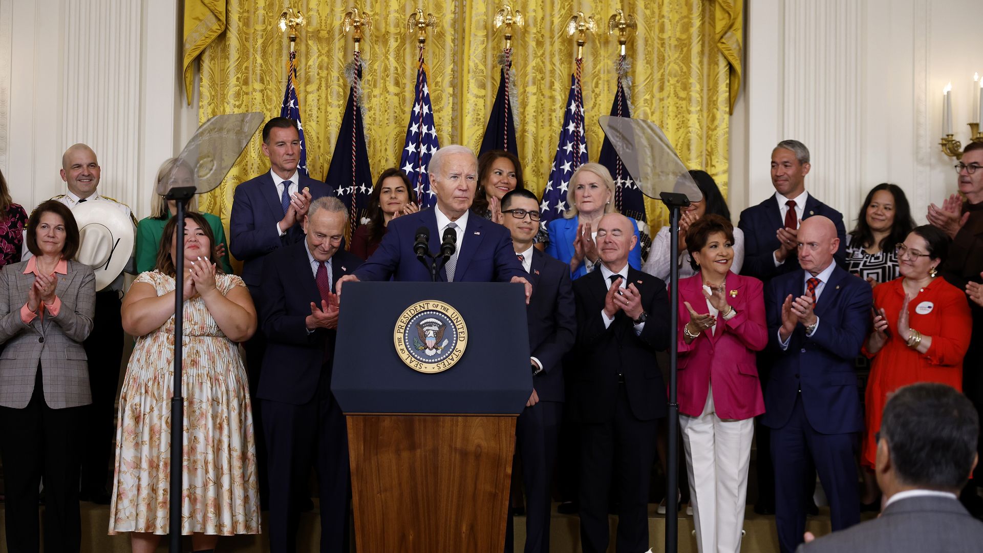 President Biden stands behind a podium at the White House surrounded by many people