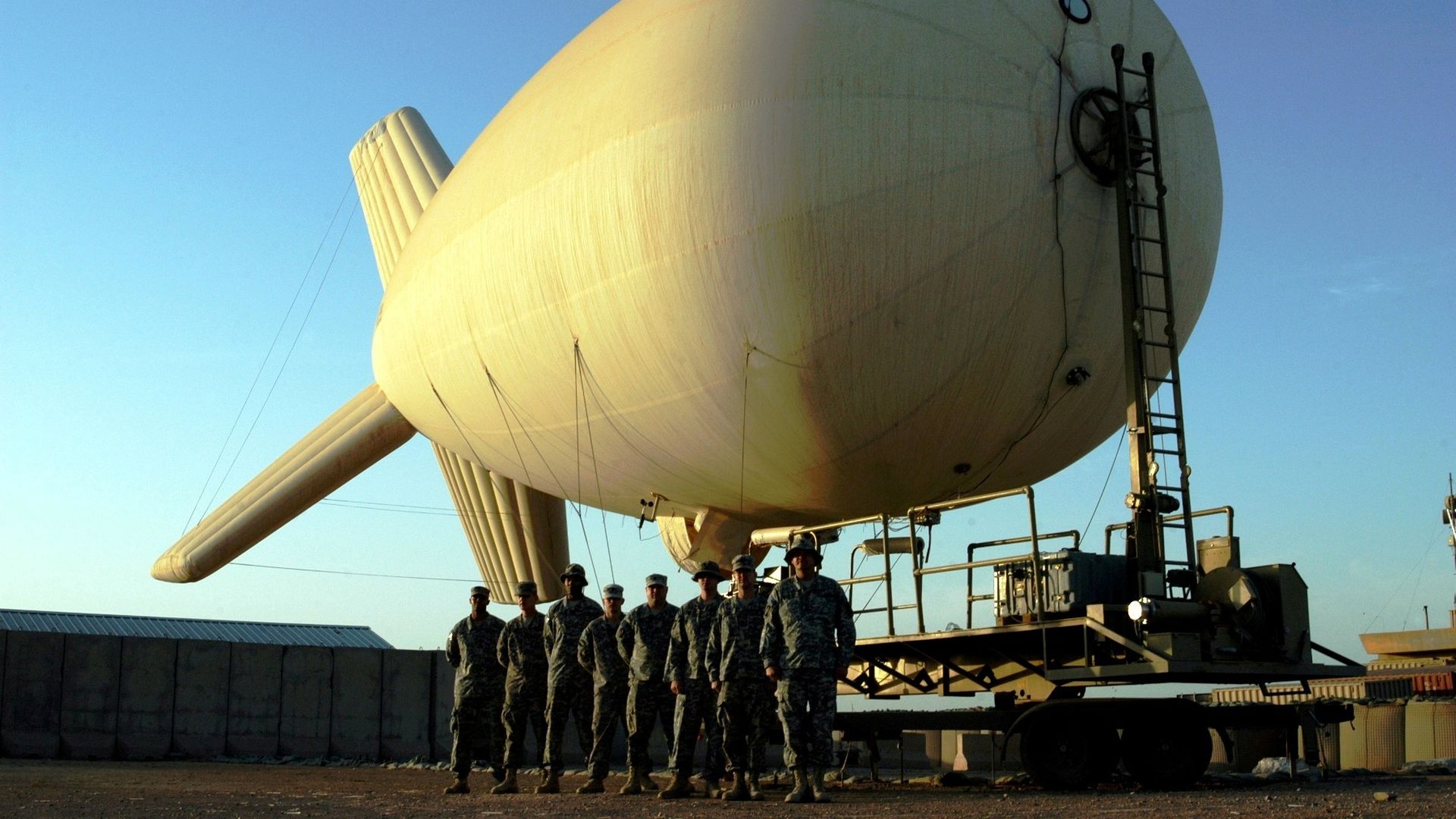 An aerostat is tethered behind a line of U.S. troops. The sky in the background is a light blue.