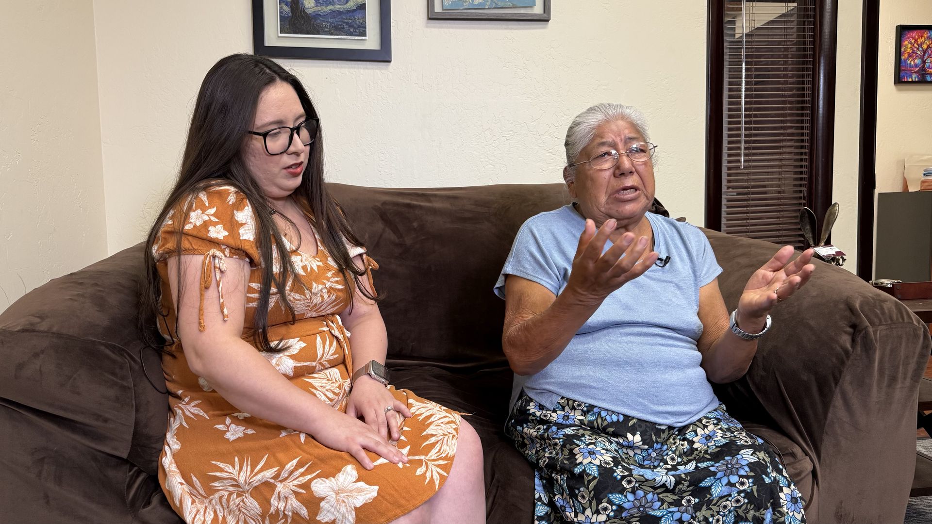 Two women sit on a brown couch in a living room: a younger woman with long dark hair and glasses in an orange floral dress, and an older woman in a light blue shirt gesturing while speaking.