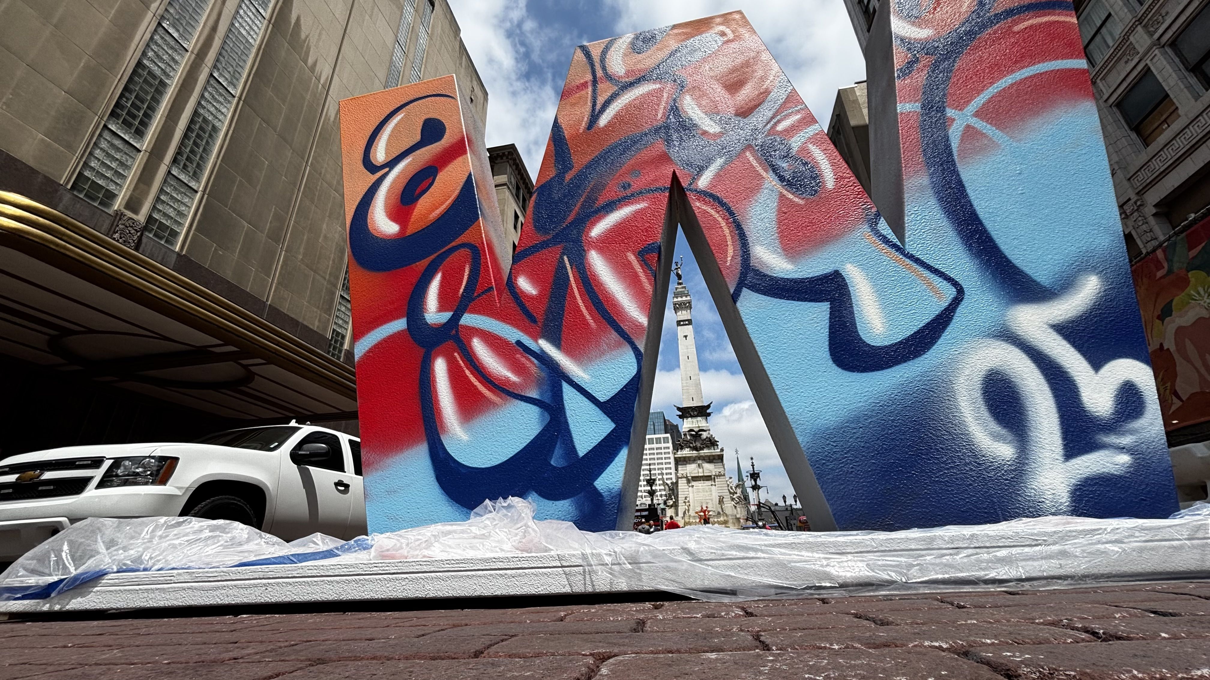 Colorful graffiti-covered large letters form an arch with a tall monument visible through the middle, set on a brick street with buildings and a white truck nearby under a partly cloudy sky.