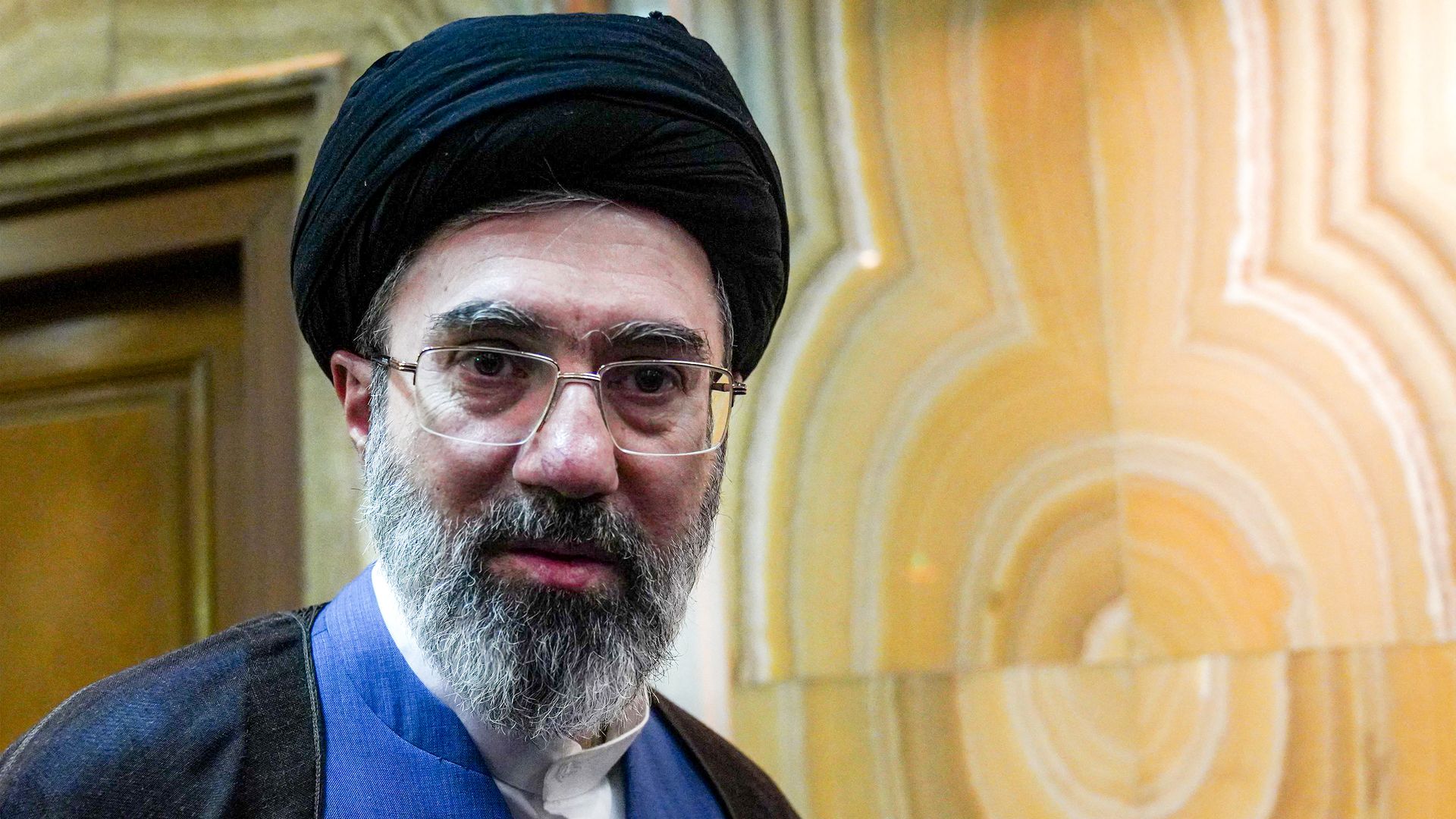 Portrait of a man with a black turban, glasses, and gray beard, wearing a blue shirt and dark robe, indoors against a warm beige patterned wall.