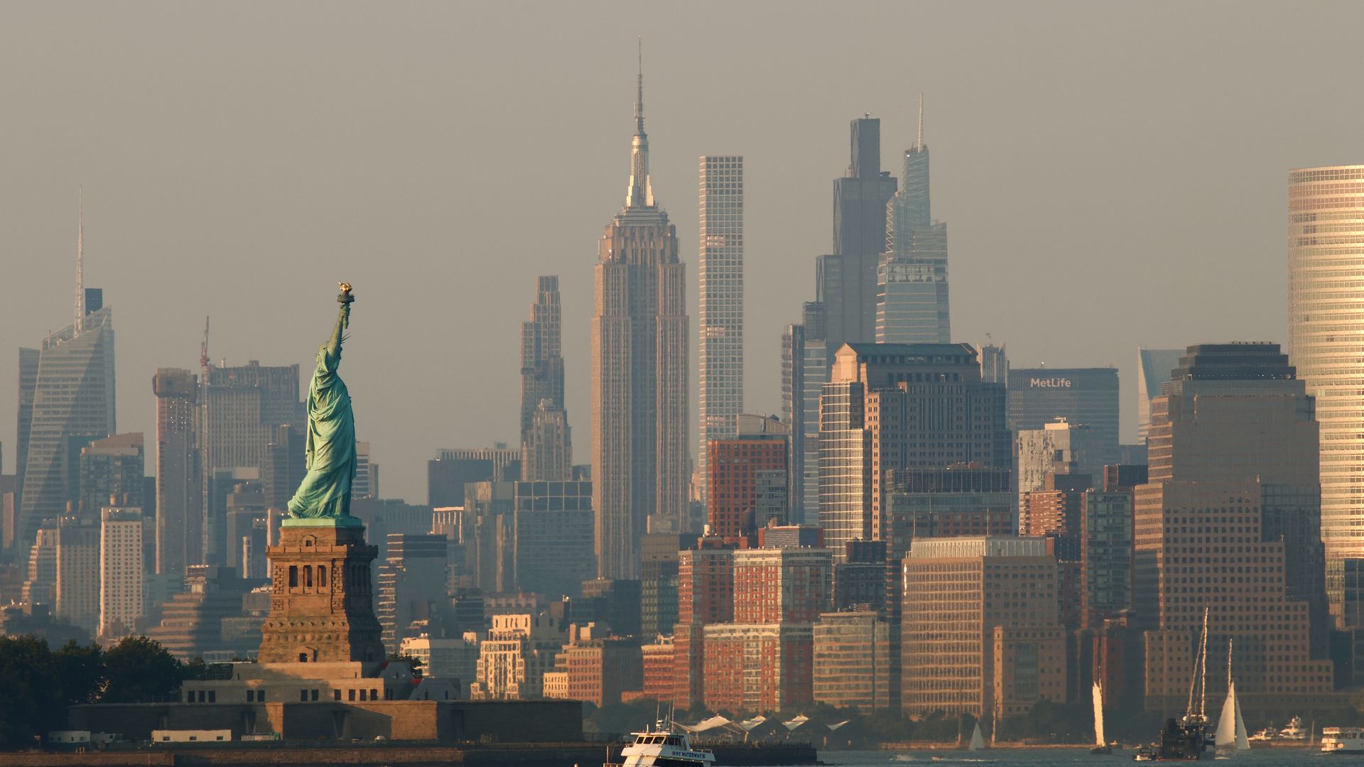 View of the Statue of Liberty with the New York City skyline in the background during a clear day, featuring boats on the water.
