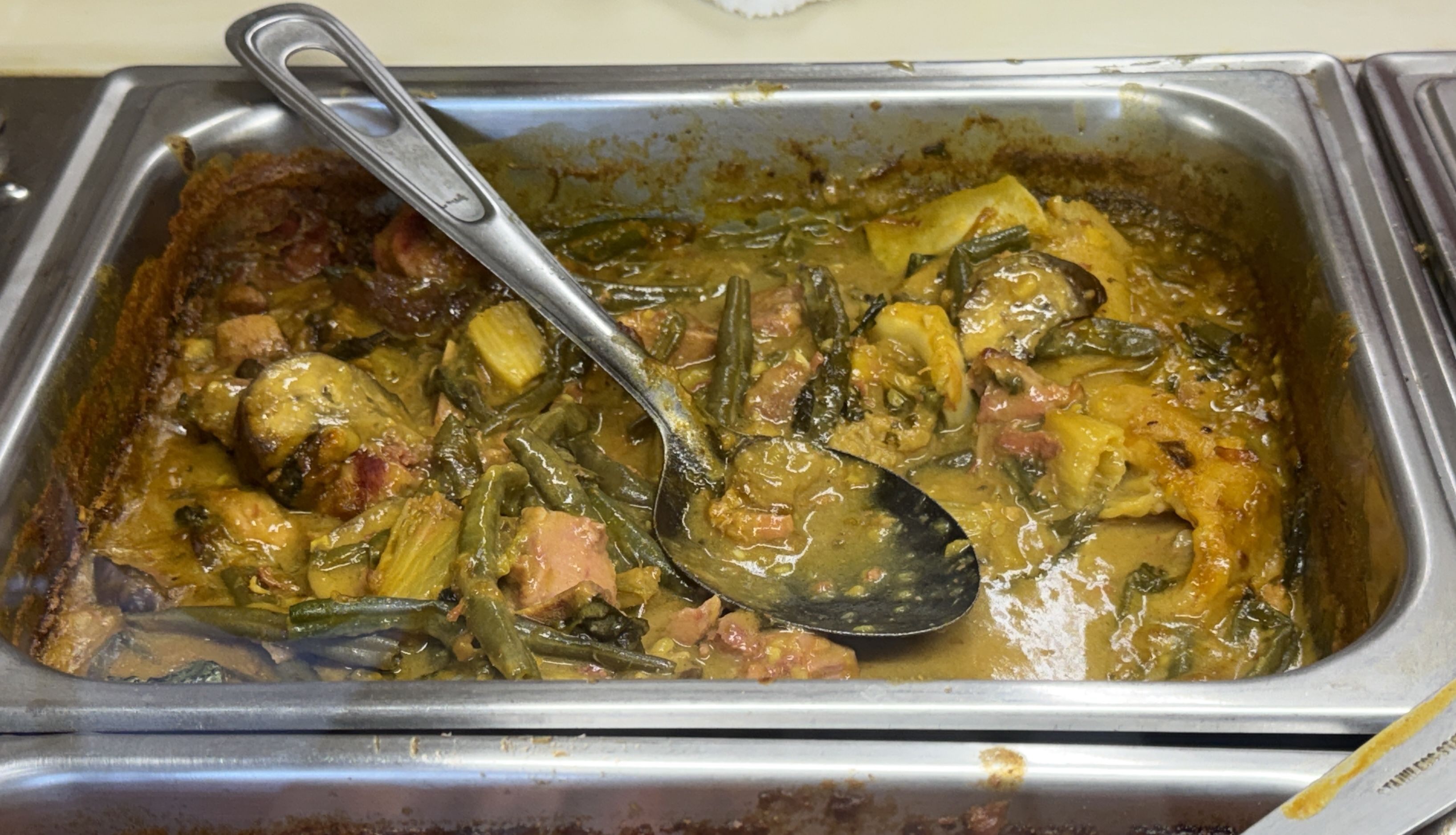 A stainless steel chafing dish filled with a chunky stew of potatoes, green beans, and meat in a yellow-brown sauce; a large metal serving spoon rests across the dish.