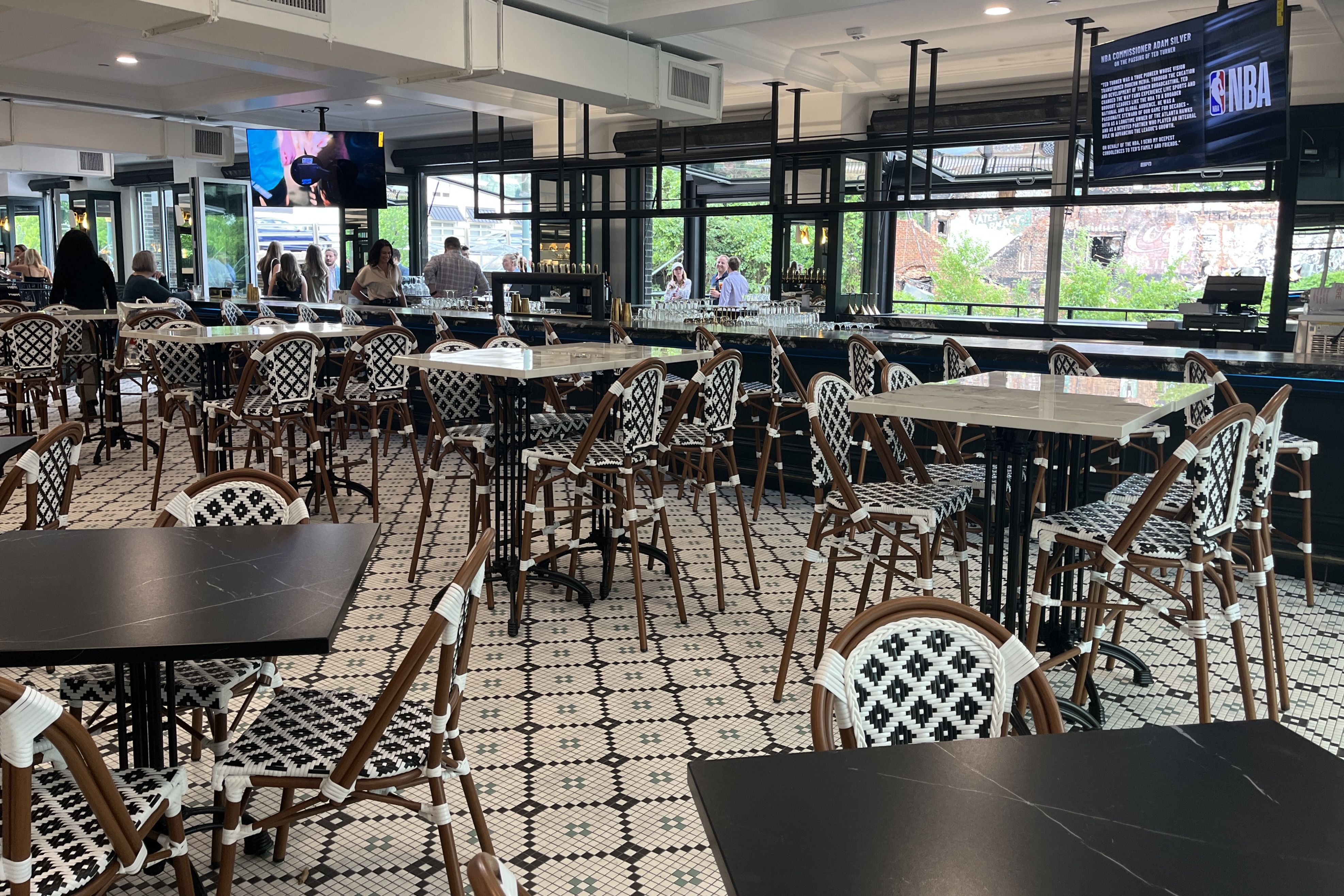 Bright indoor bar/restaurant with black-and-white patterned wicker chairs, white tabletops, and a long counter. Patrons stand by the bar; large windows overlook greenery and a brick building. Two TVs show NBA.