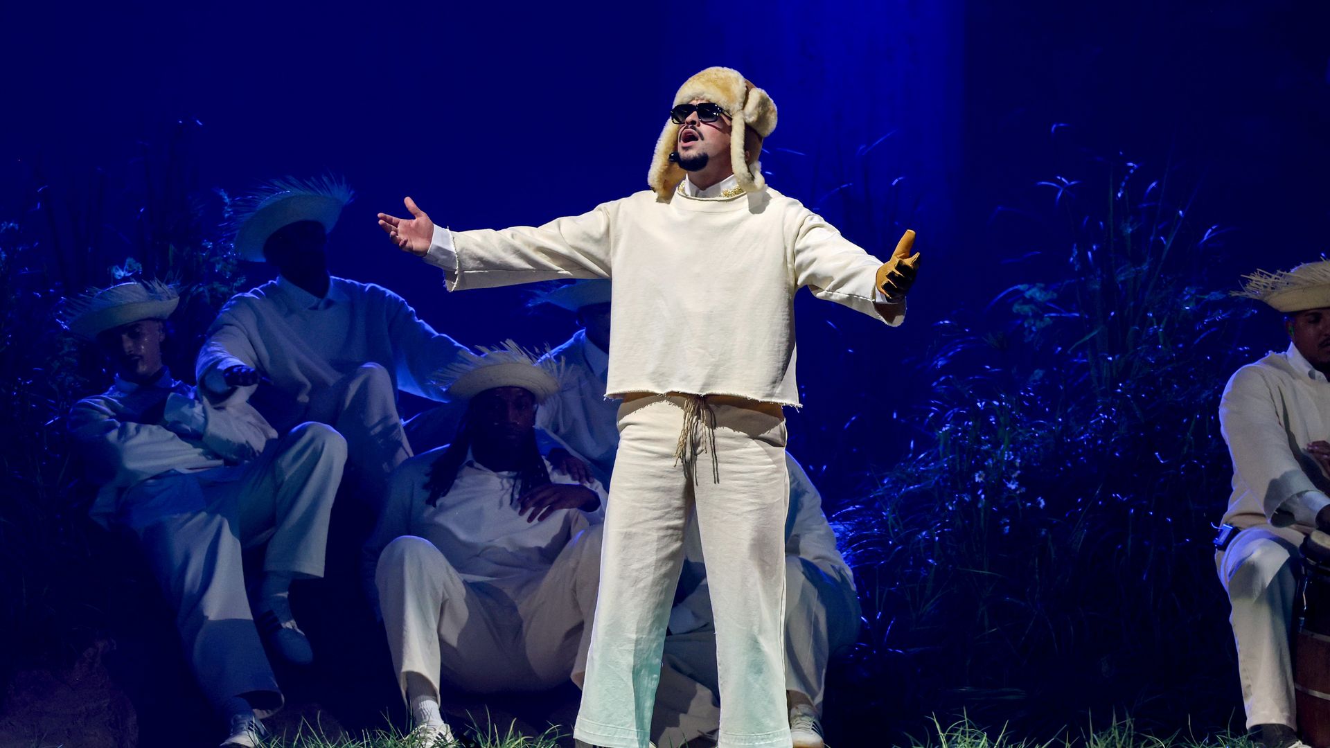 Bad Bunny, wearing a cream fur hat and dark glasses and dressed in a cream top and pants with white sneakers, gestures as he performs on stage.