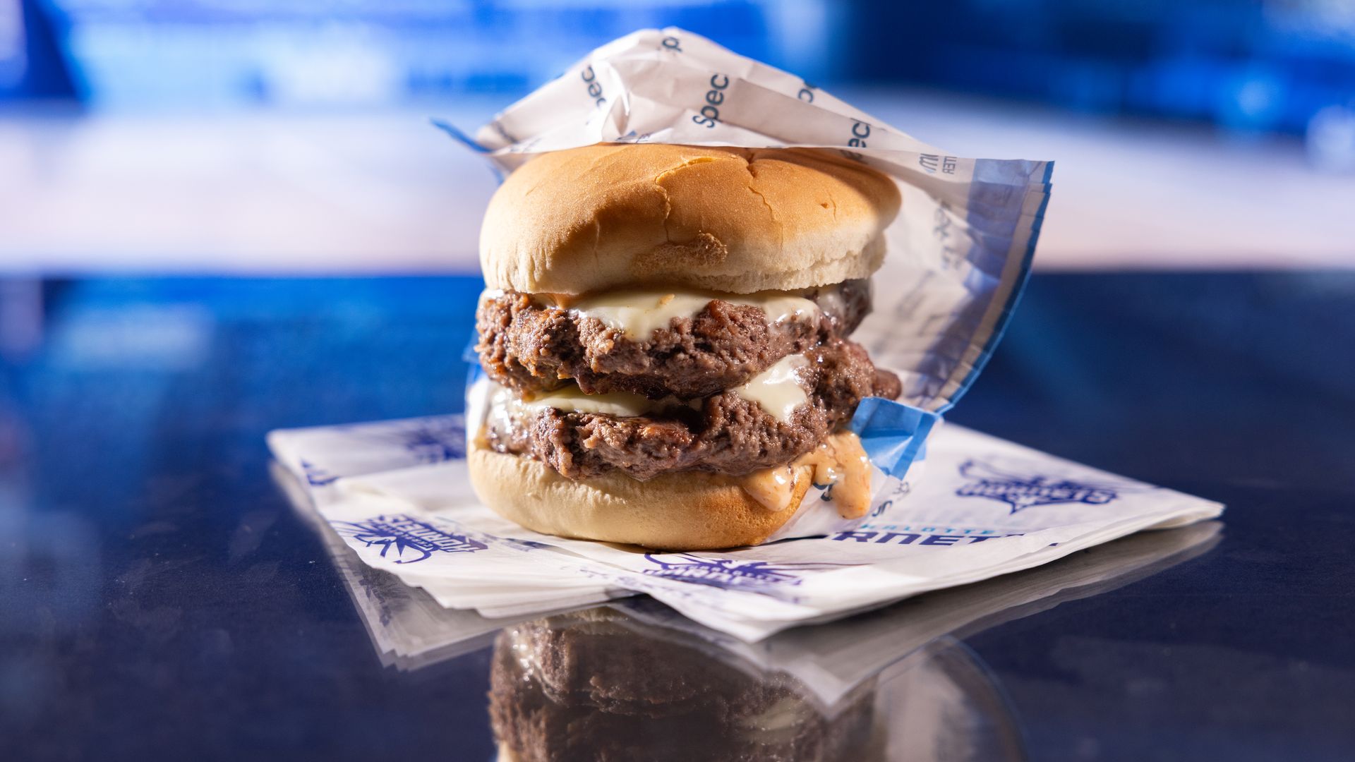 Double cheeseburger with melted cheese and sauce dripping from a soft bun, placed on branded wax paper on a reflective surface, with a blue blurred background.