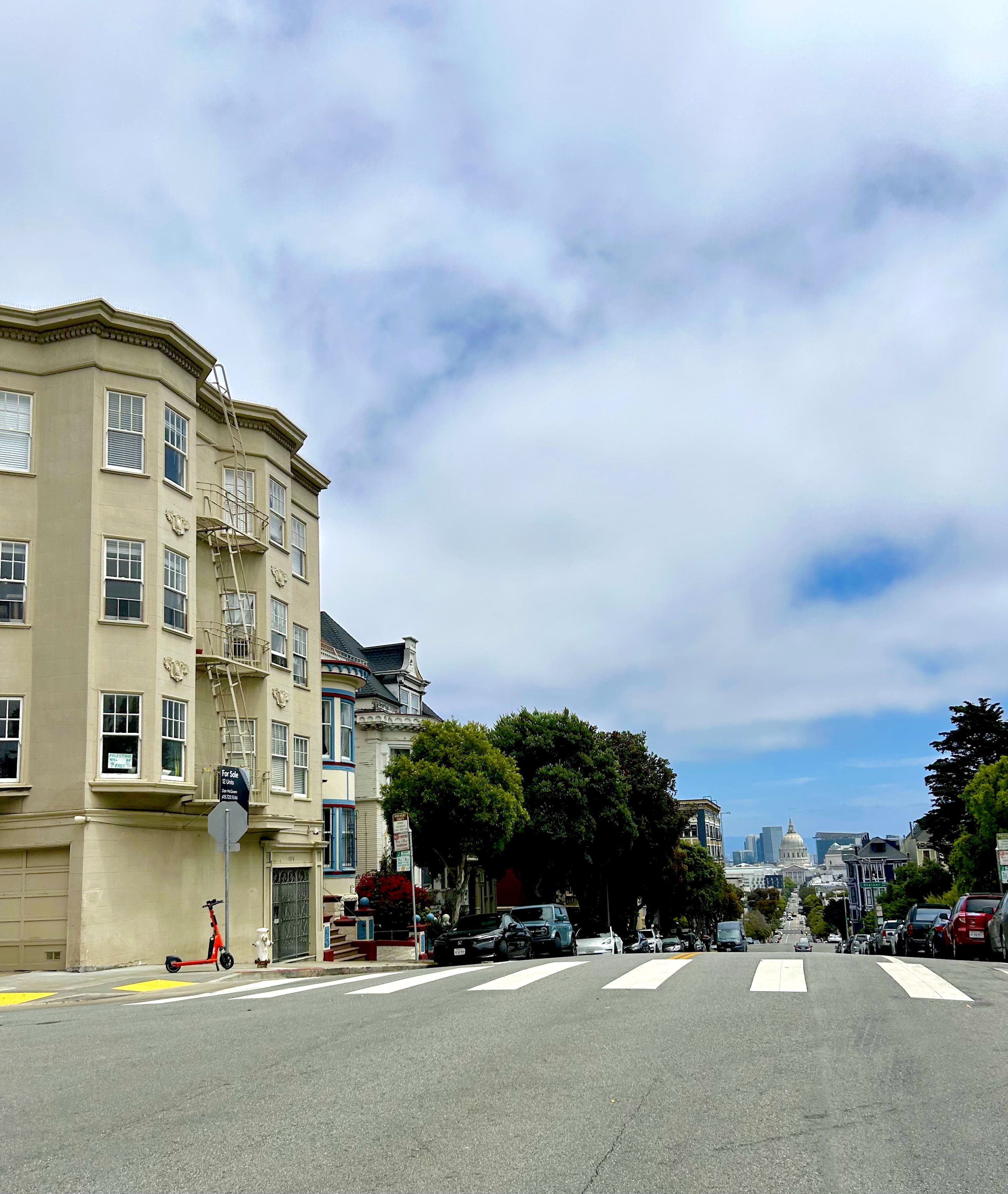 View down a steep city street with crosswalk and parked cars, beige and colorful buildings on left, green trees, and a domed government building in distance under a cloudy blue sky.