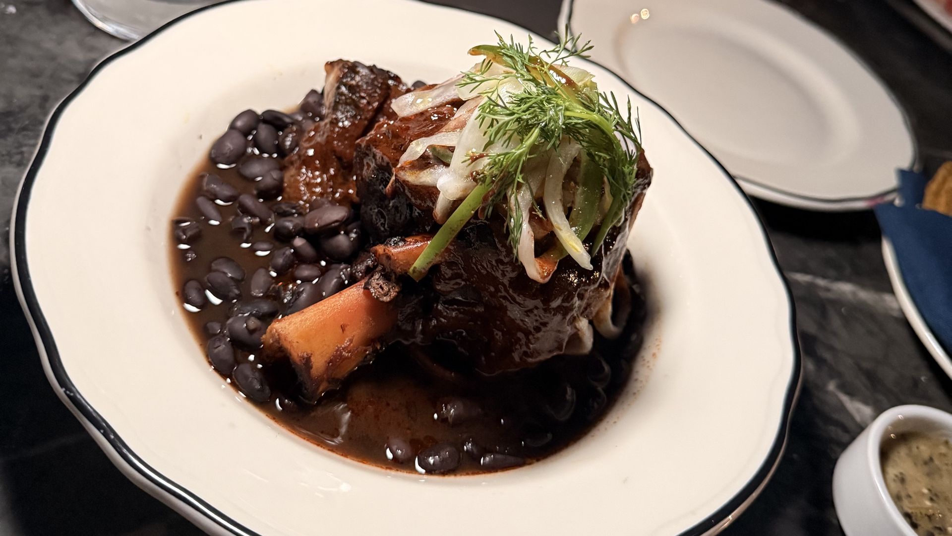 Plate of braised meat with dark sauce, black beans, and carrot chunks, topped with greens; a glass of water sits on a dark marble table, with two diners blurred in the background.