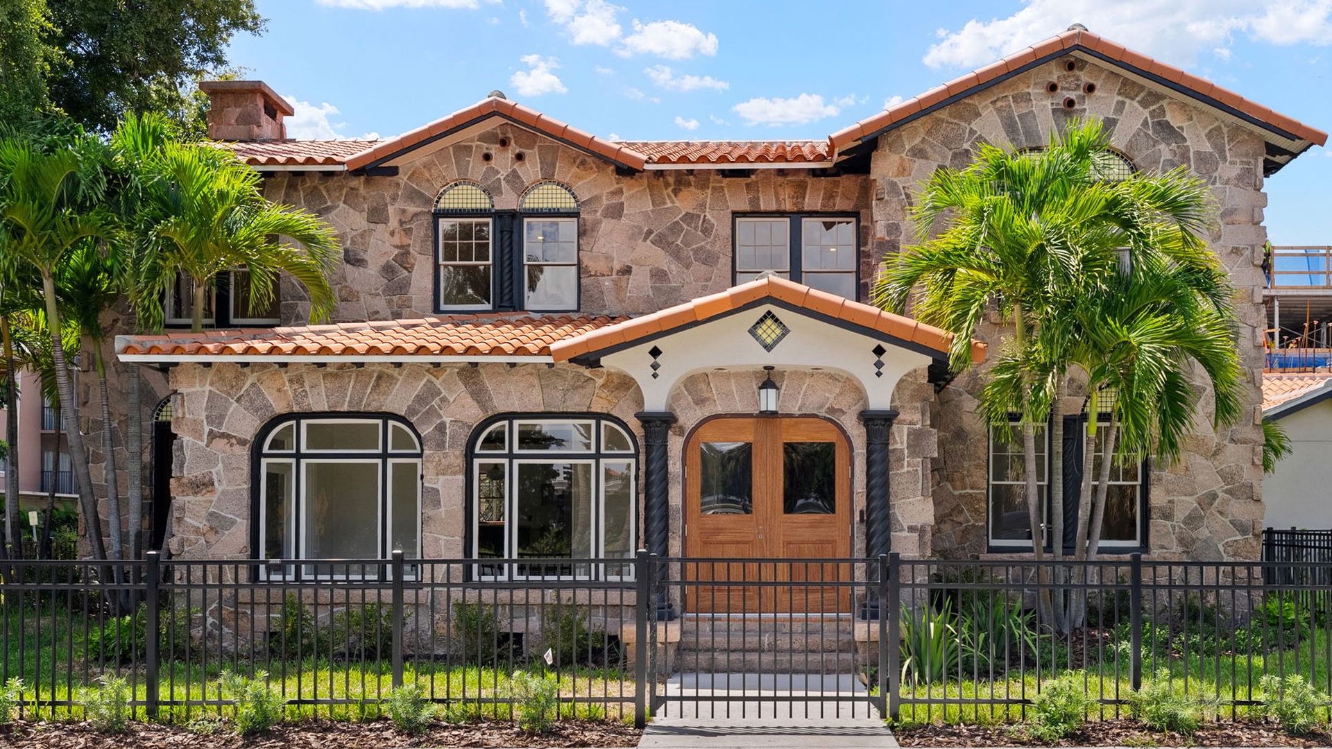 Two-story stone house with a red clay tile roof, arched windows, and a wooden double door under a small portico. Palm trees flank the front; a black wrought-iron fence runs along the yard.
