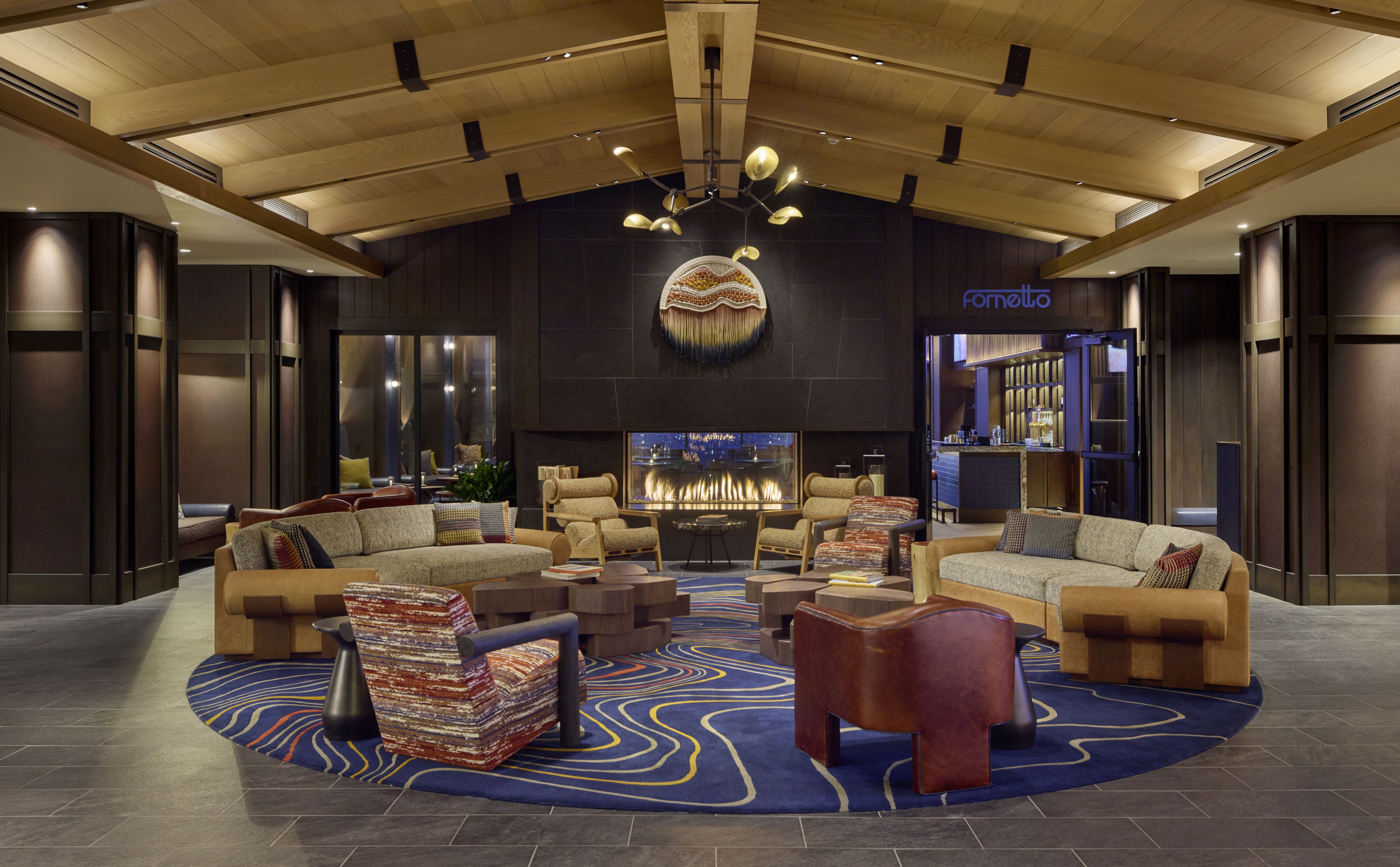 Luxurious hotel lobby lounge with a vaulted wooden ceiling, modern chandelier, and dark stone fireplace. Beige sofas and colorful chairs surround a blue patterned rug; a blue Fornetto sign sits near the bar.