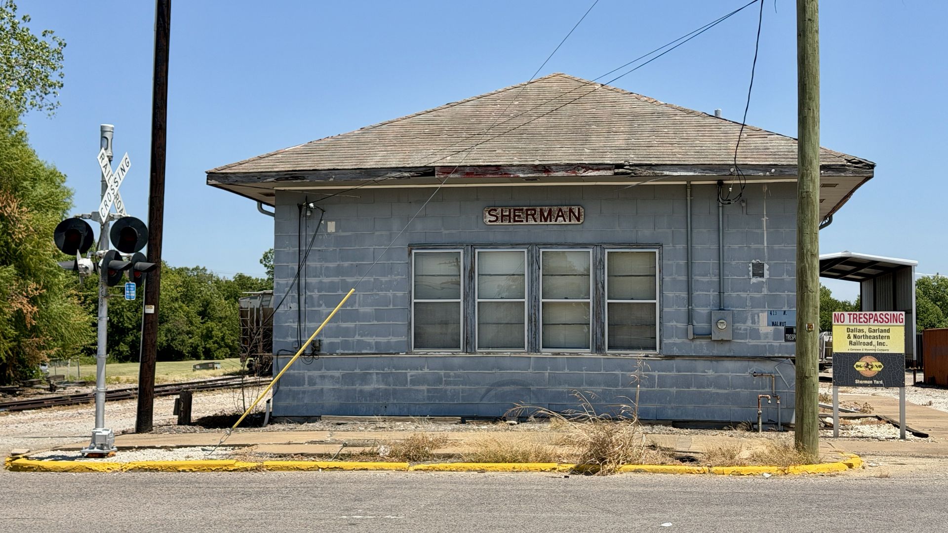 Old light-blue brick railroad station with a peaked roof labeled "SHERMAN", three windows, nearby crossing signals, utility wires, a yellow curb, and a "NO TRESPASSING" sign.