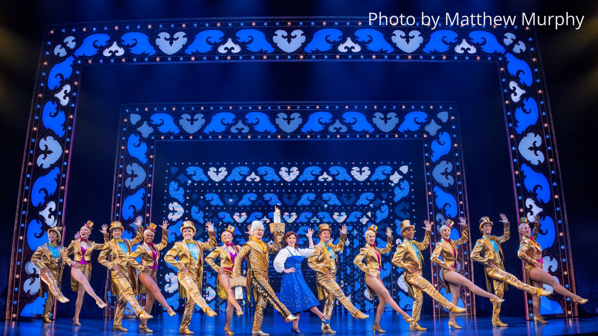 Stage musical: dancers in gold sequined suits and top hats kick in unison, flanking a man in gold and a woman in a blue dress, against an ornate blue backdrop with heart motifs.