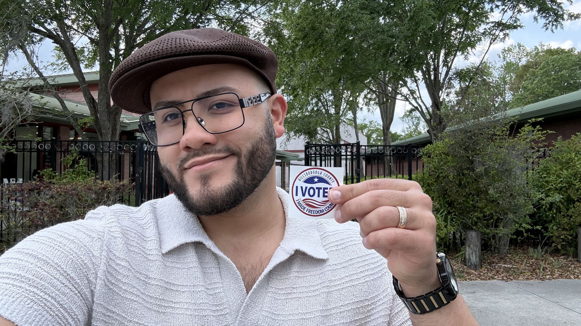A man with a brown flat cap, glasses, and a trimmed beard smiles at the camera while holding a circular "I VOTE" sticker in red, white, and blue; trees and a fence are in the background.