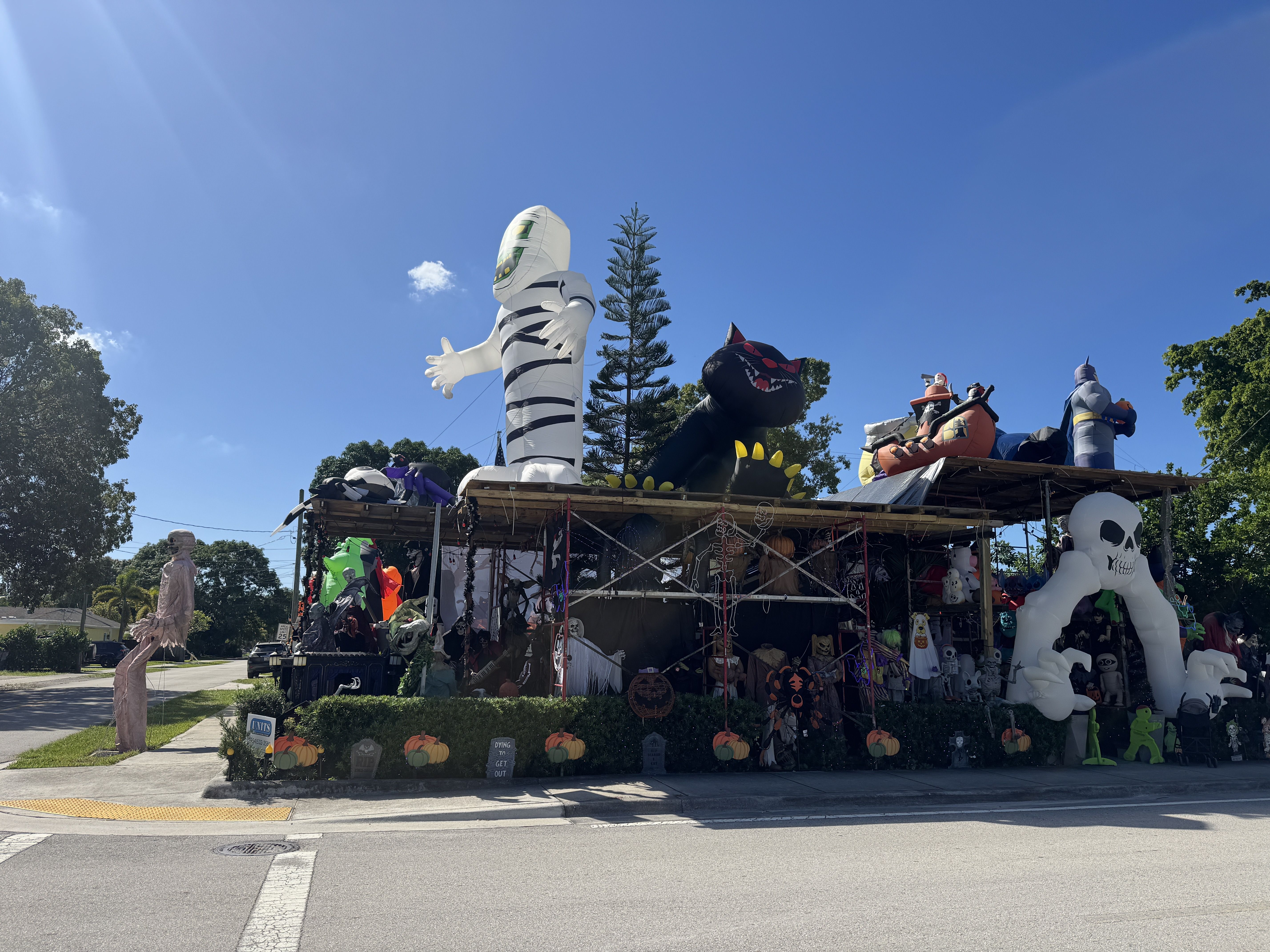Blue sky over a Halloween display featuring tall inflatables: a white striped mummy, a black dragon, plus skeletons, pumpkins, and other spooky figures on a storefront booth.